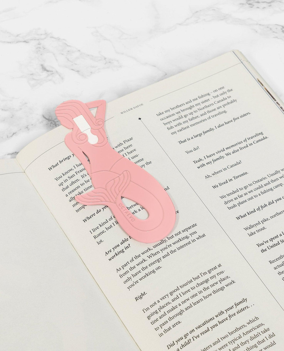 The pink OCTAEVO brass Bookmark Sirena rests on an open book displayed on a marble surface.