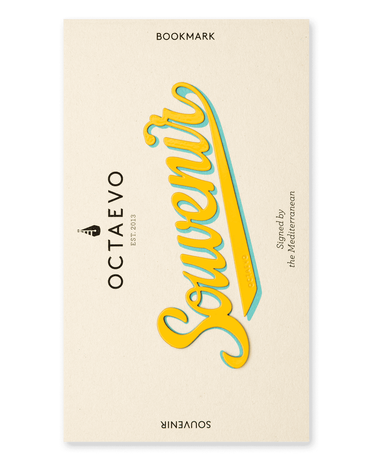 The brass Bookmark Souvenir by OCTAEVO features yellow script on a cream background with additional small text.