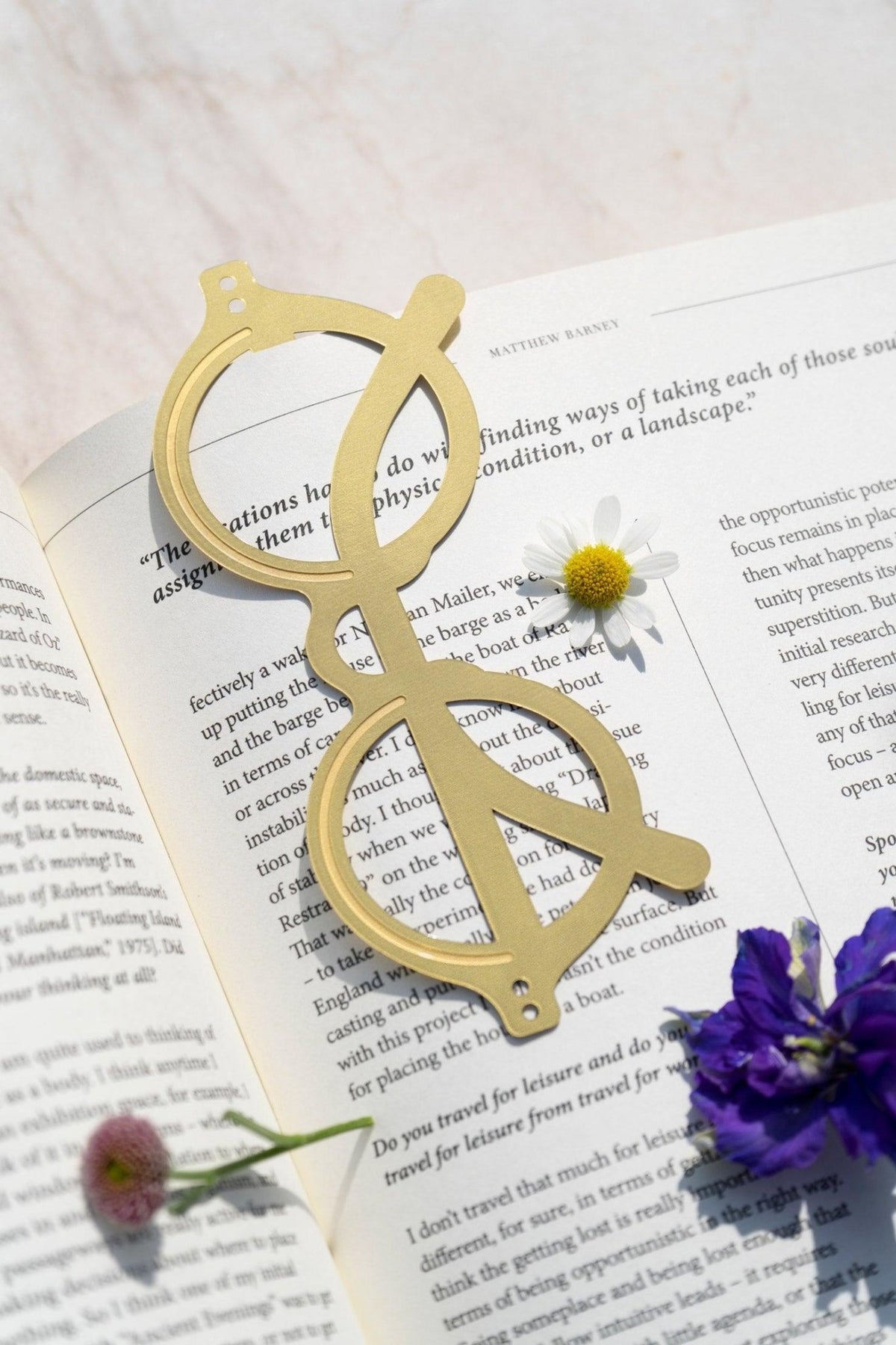 The Voyage Bookmark by OCTAEVO—brass sunglasses-shaped, round frames—rests on an open book with small flowers nearby.
