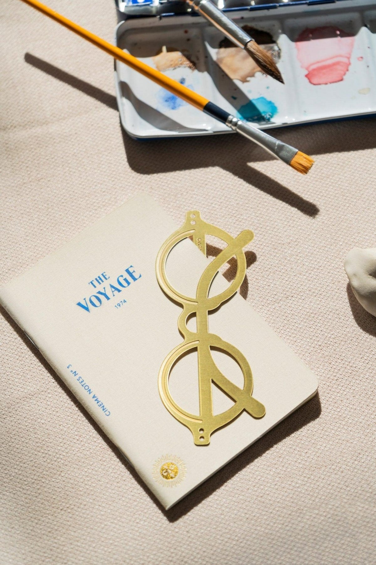 A paintbrush and palette beside OCTAEVO notebook The Voyage, featuring a gold sunglass bookmark on the cover.
