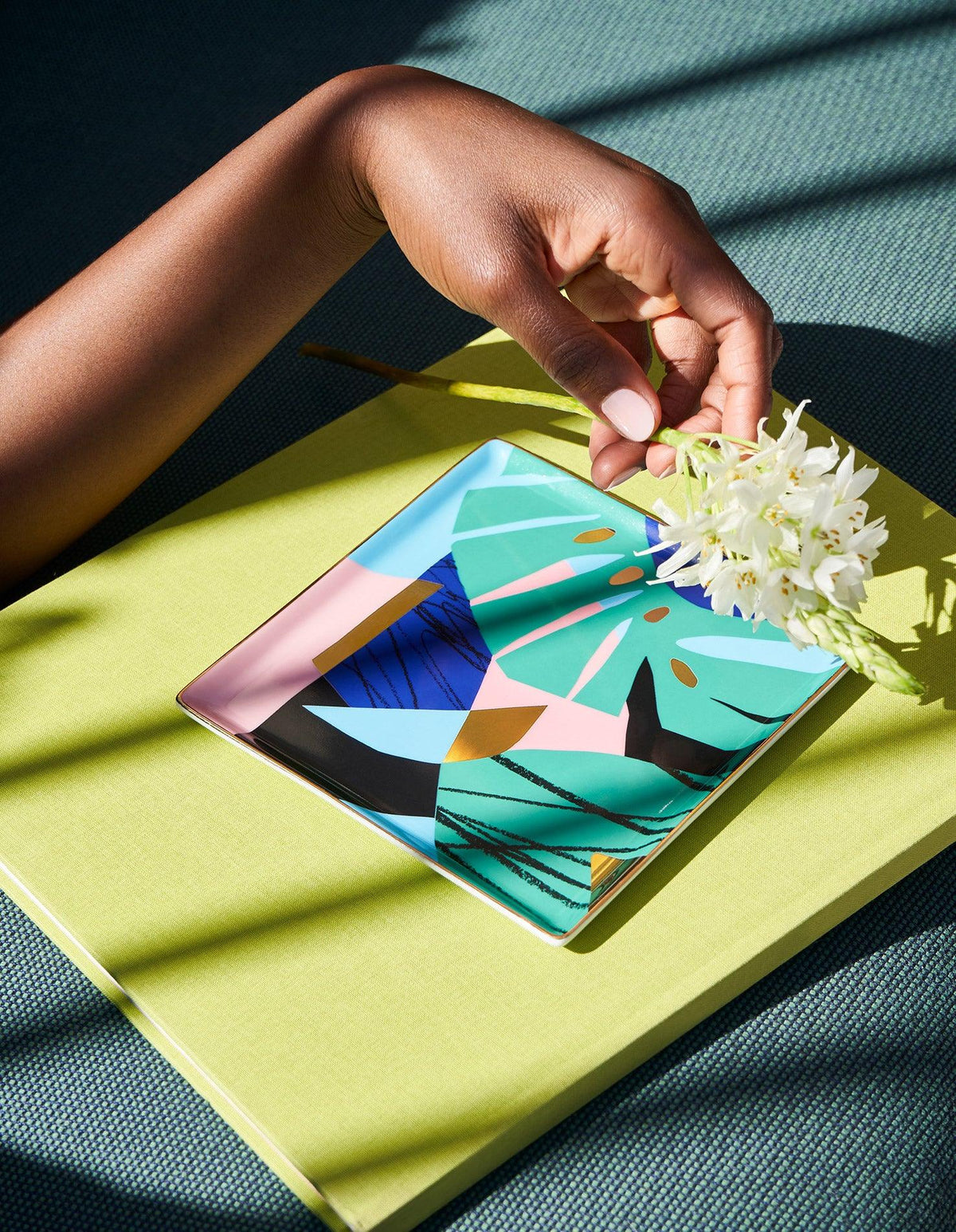 A hand holds white flowers above the Capri Ceramic Tray by OCTAEVO on a lime green surface.