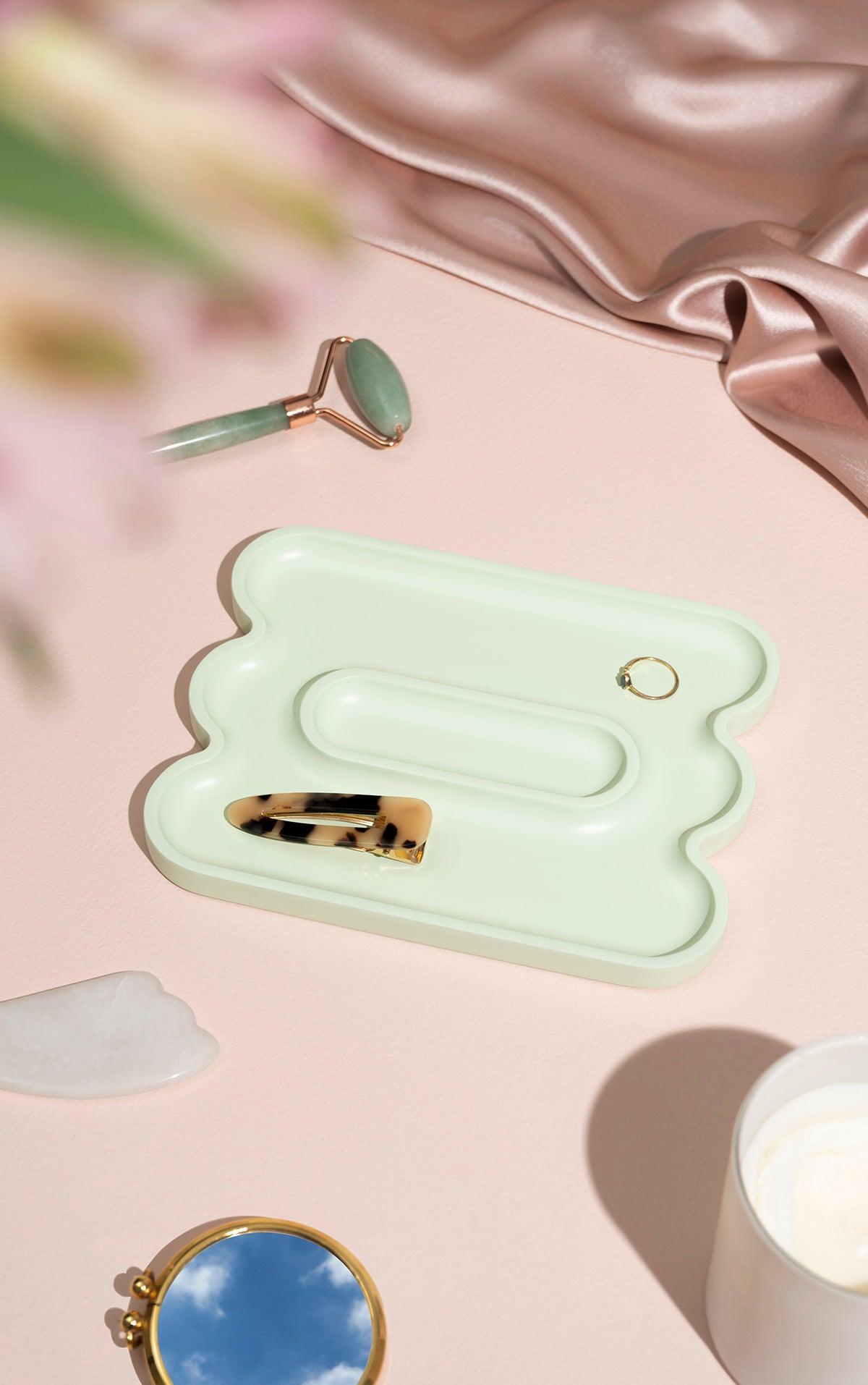Templo Wave Catchall by OCTAEVO holds a hair clip and ring, beside a jade roller, mirror, cream, and pink satin.
