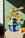A hand arranges yellow and white flowers in a colorful face-shaped blue vase on a table with fruits. 