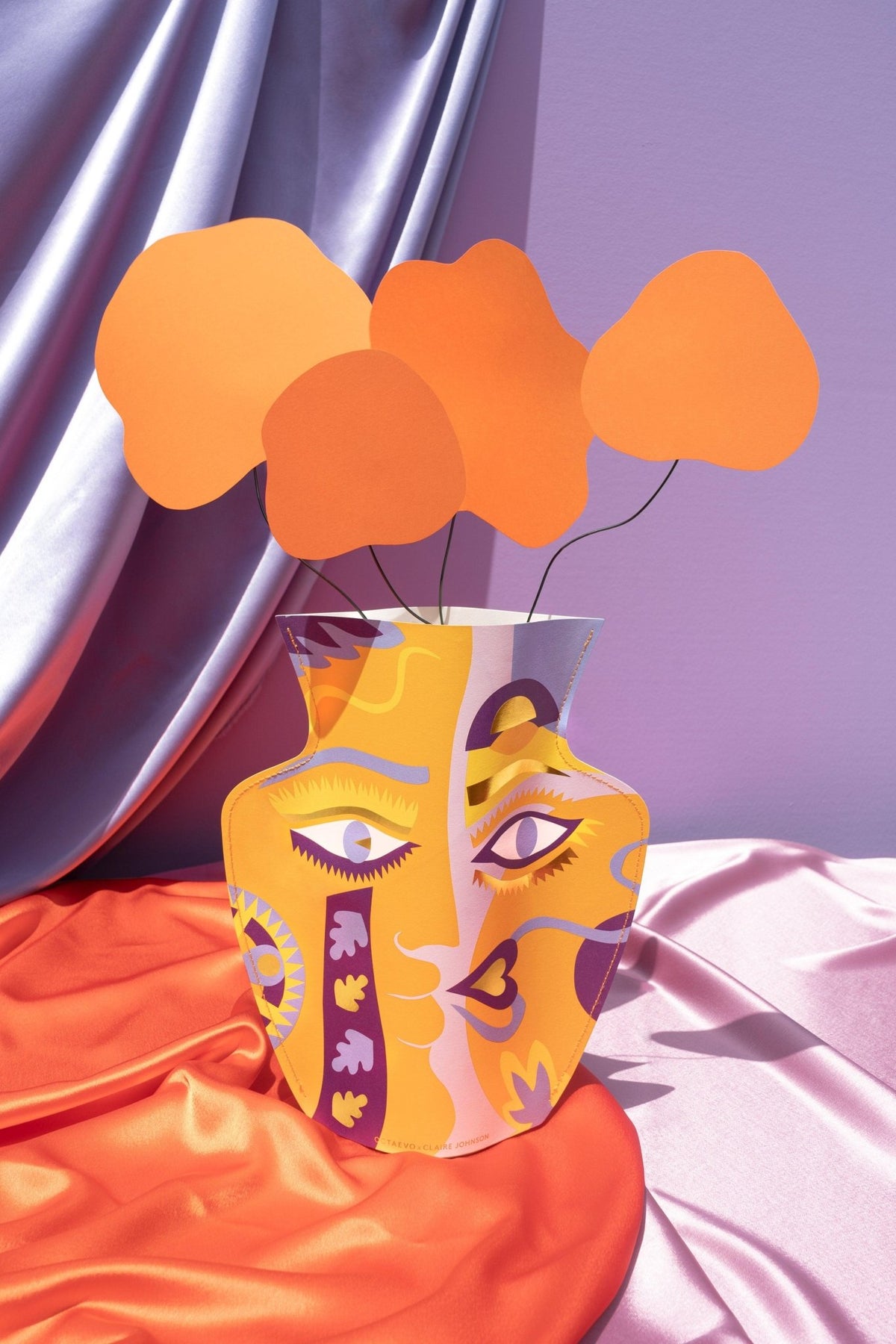 The orange Octaevo Claire Johnson Paper Vase with a face design holds paper flowers on draped satin fabric.