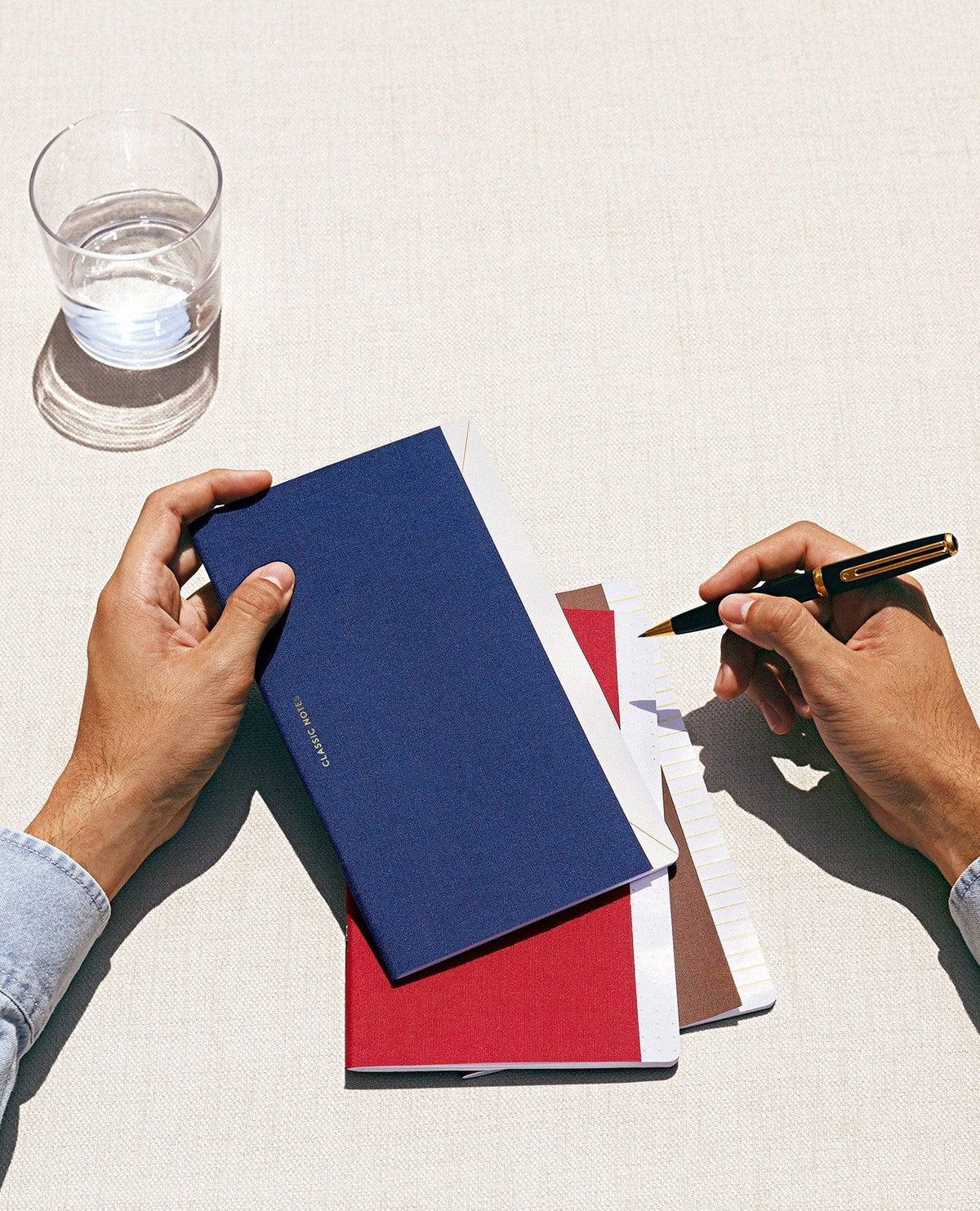 A person holds an blue OCTAEVO Classic Notes notebook and pen, with stacked notebooks and water on a light table.