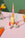 Three Octaevo Dip Dye Candles Pomegranate in wavy holders on a pink table with flowers and grapefruit nearby.