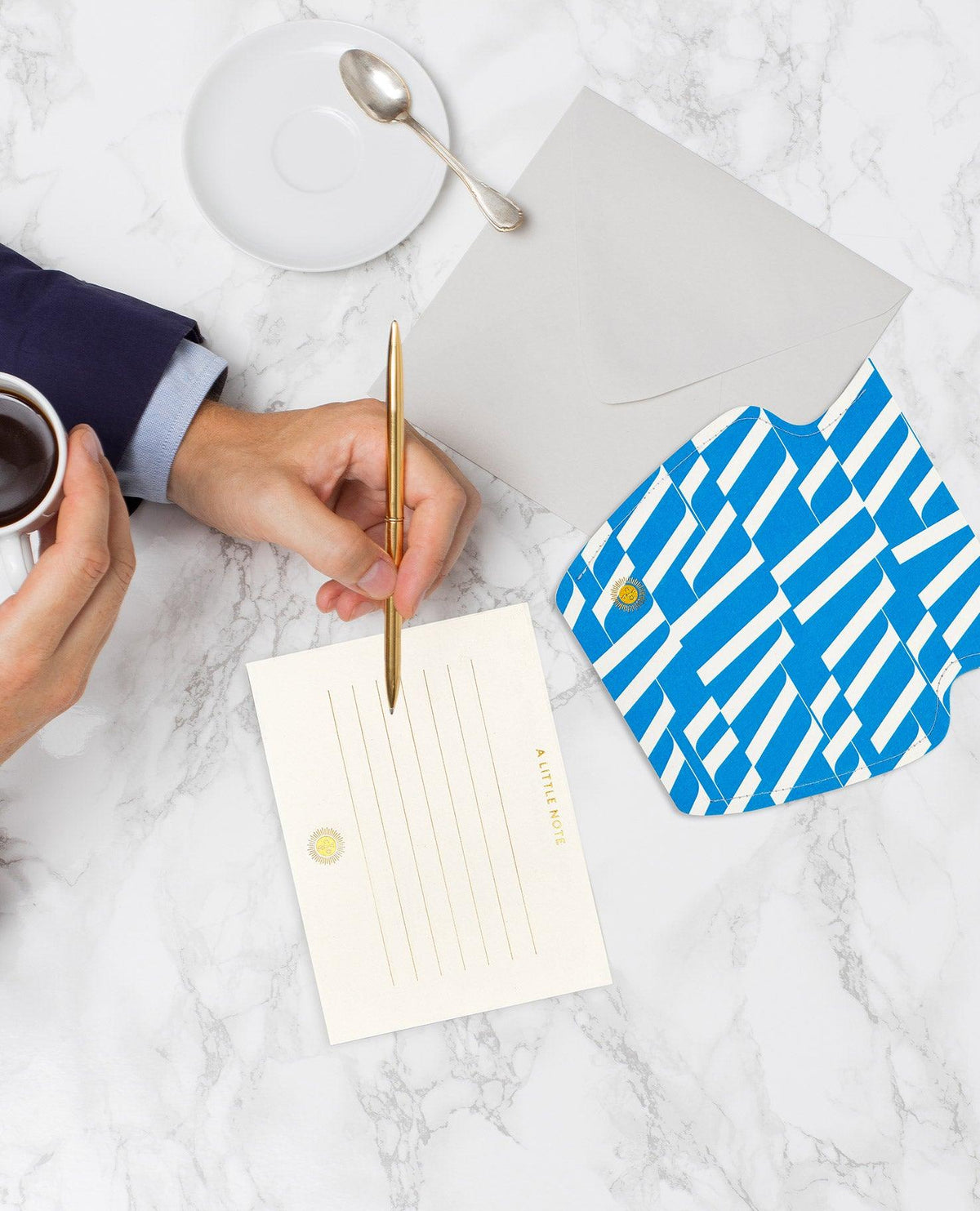 Une personne écrit sur une carte à côté du mini vase en papier Domus OCTAEVO et d'une enveloppe à rayures bleues, en sirotant un café.