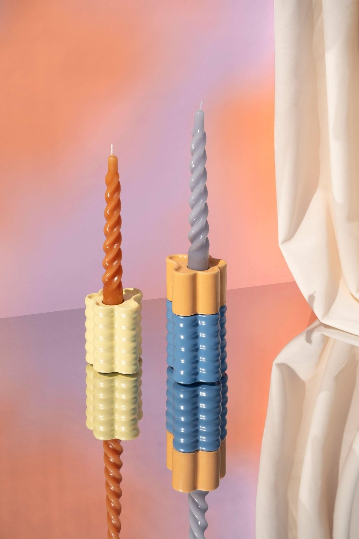 Two twisted candles in Octaevo Flora Candle Holders in orange sit on a reflective surface with a pastel gradient background.