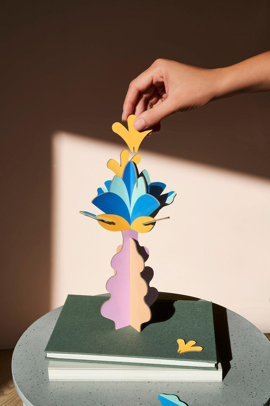 A hand adds a yellow shape to Octaevos Flower Paper Sculpture 2, glowing on books in Elysian Fields sunlight.