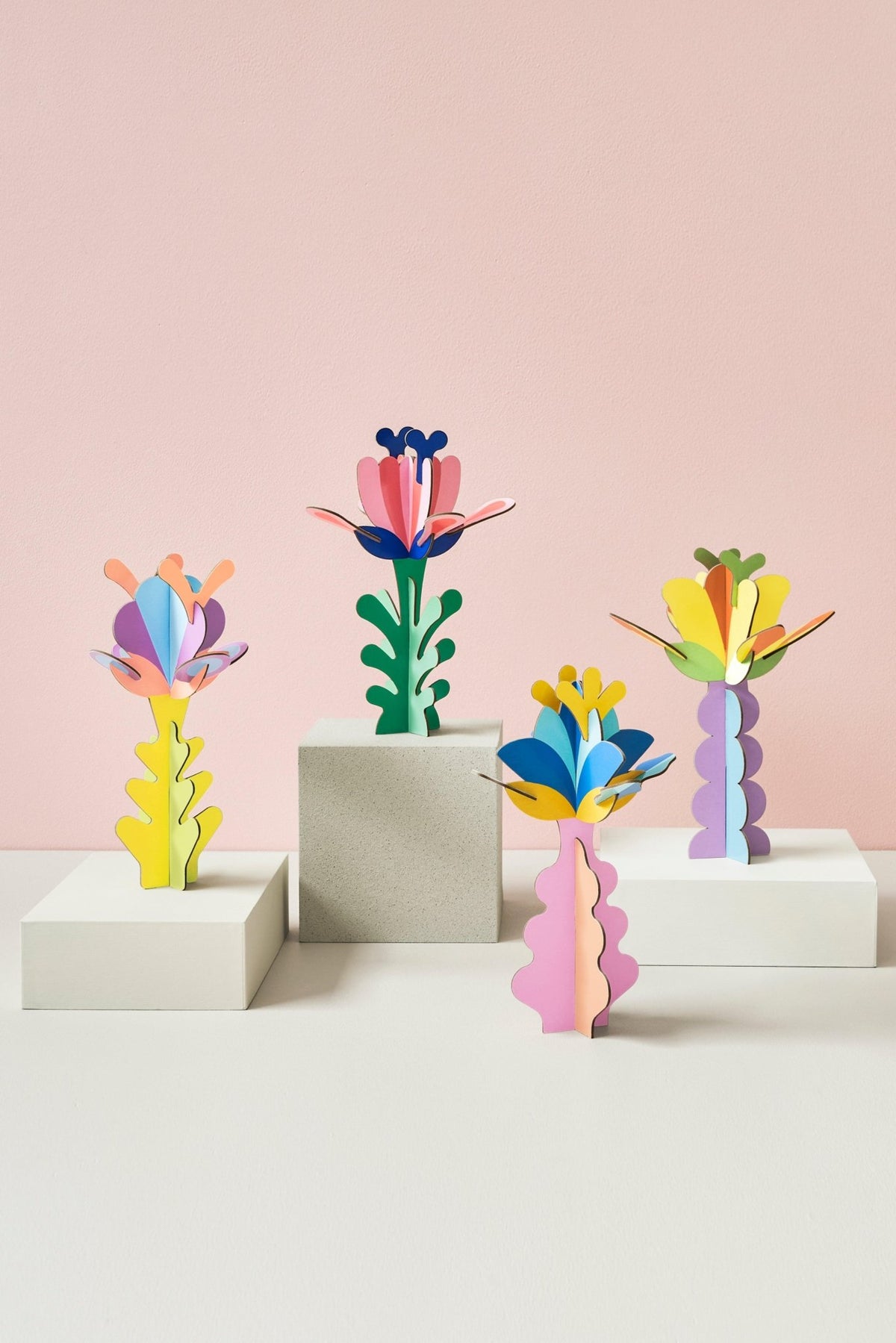 Four vibrant OCTAEVO Aster Flower Paper Sculptures stand on blocks against a pastel pink background.