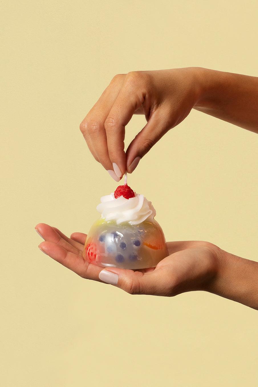 A hand holds Octaevos Fruit Jelly Dessert Candle while another places a cherry on top, creating cute tabletop decor.