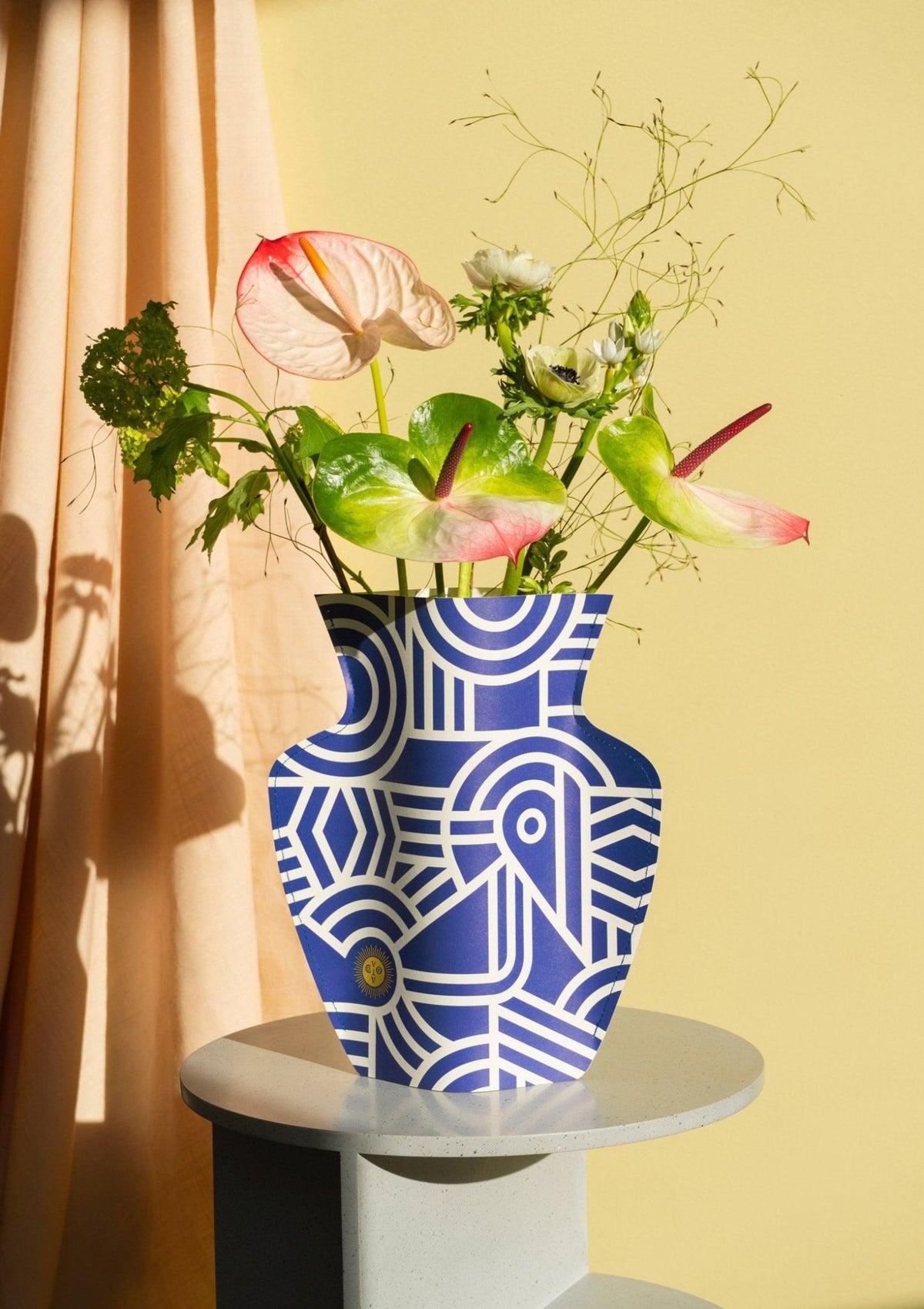 The Greco Paper Vase by OCTAEVO with assorted flowers sits on a round table against a yellow background.