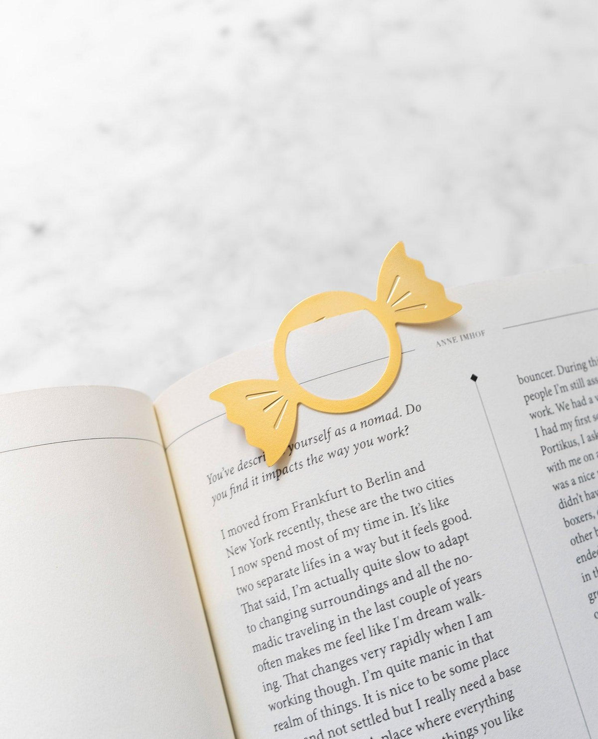 A candy-shaped gold brass bookmark by OCTAEVO rests on an open book with text over marble. 