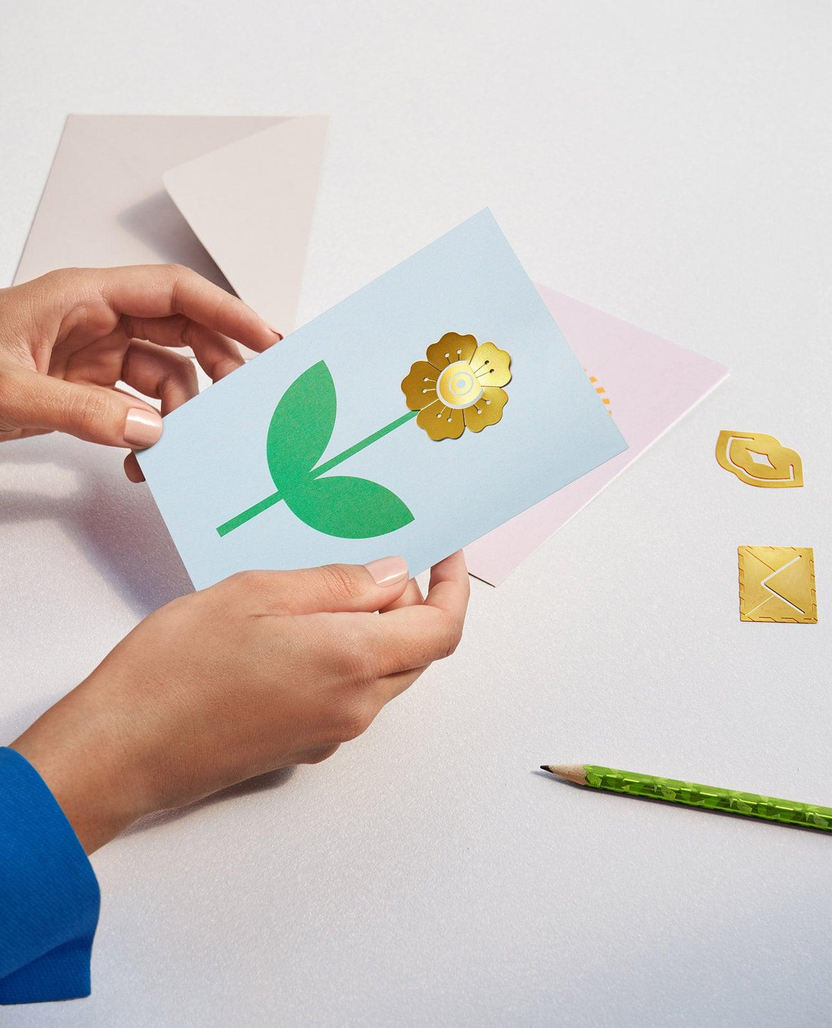 Hands hold the OCTAEVO Flower Greeting Card with envelope, pencil, and gold stickers on a white surface.