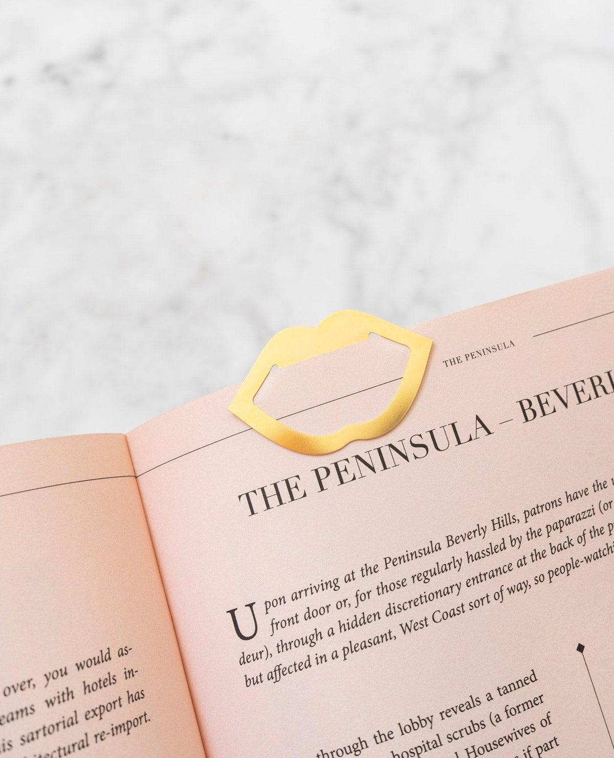 Gold, lips-shaped metal clip bookmark on pink book beside OCTAEVO Greeting Card Kiss, on a marble surface.