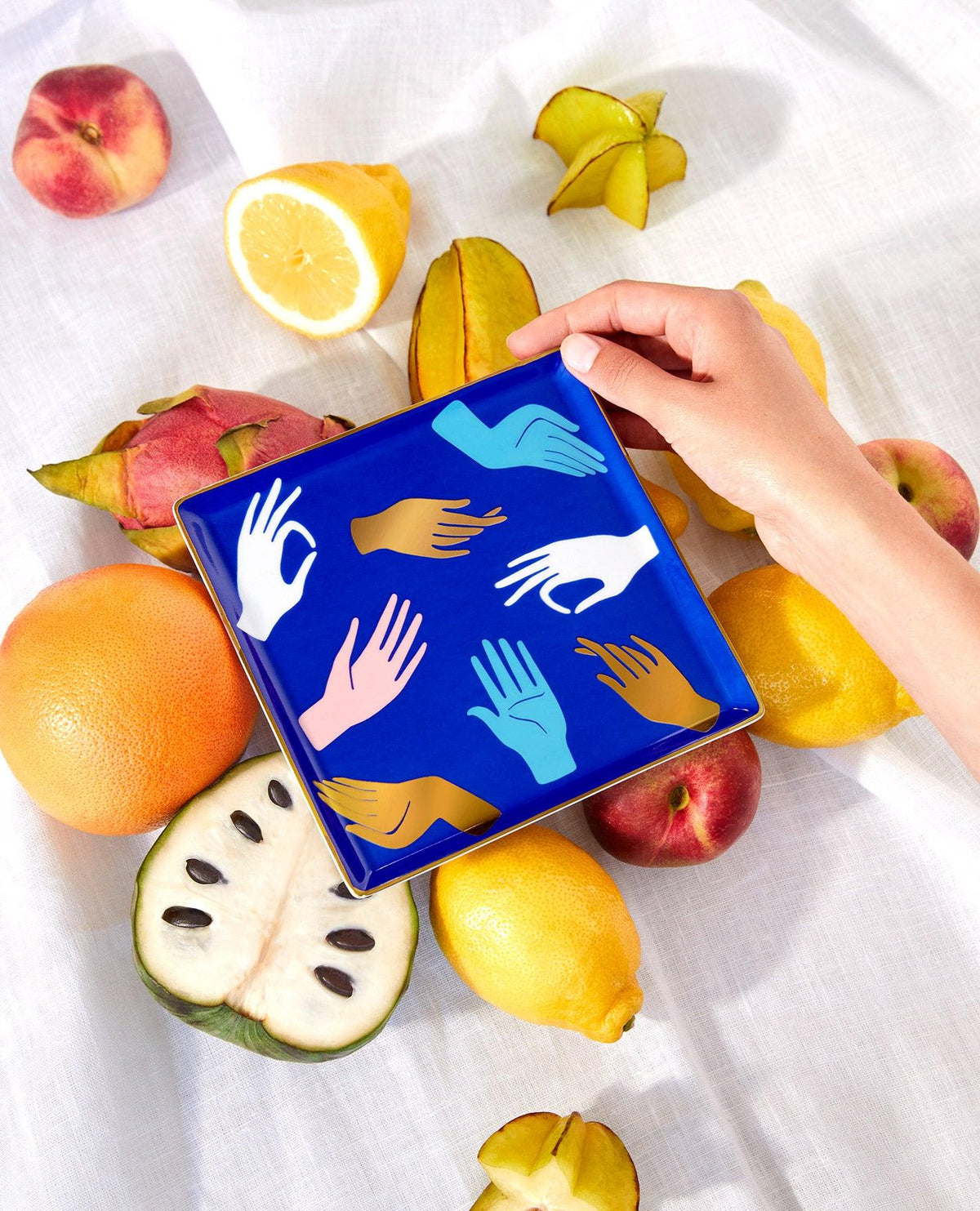 A hand holds the blue OCTAEVO Hamsa Ceramic Tray with colorful prints, placed on fruits on a white cloth.