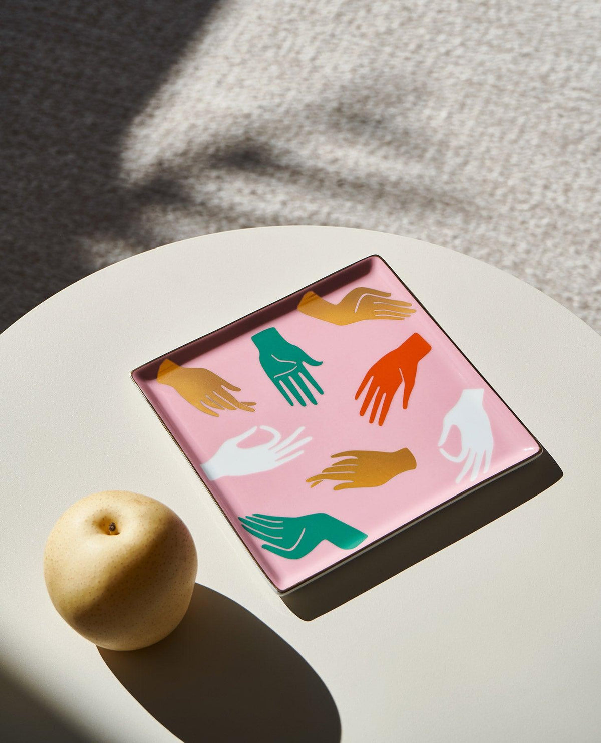 The OCTAEVO Hamsa Ceramic Tray, square and pink with colorful designs, sits on a table beside a round fruit.