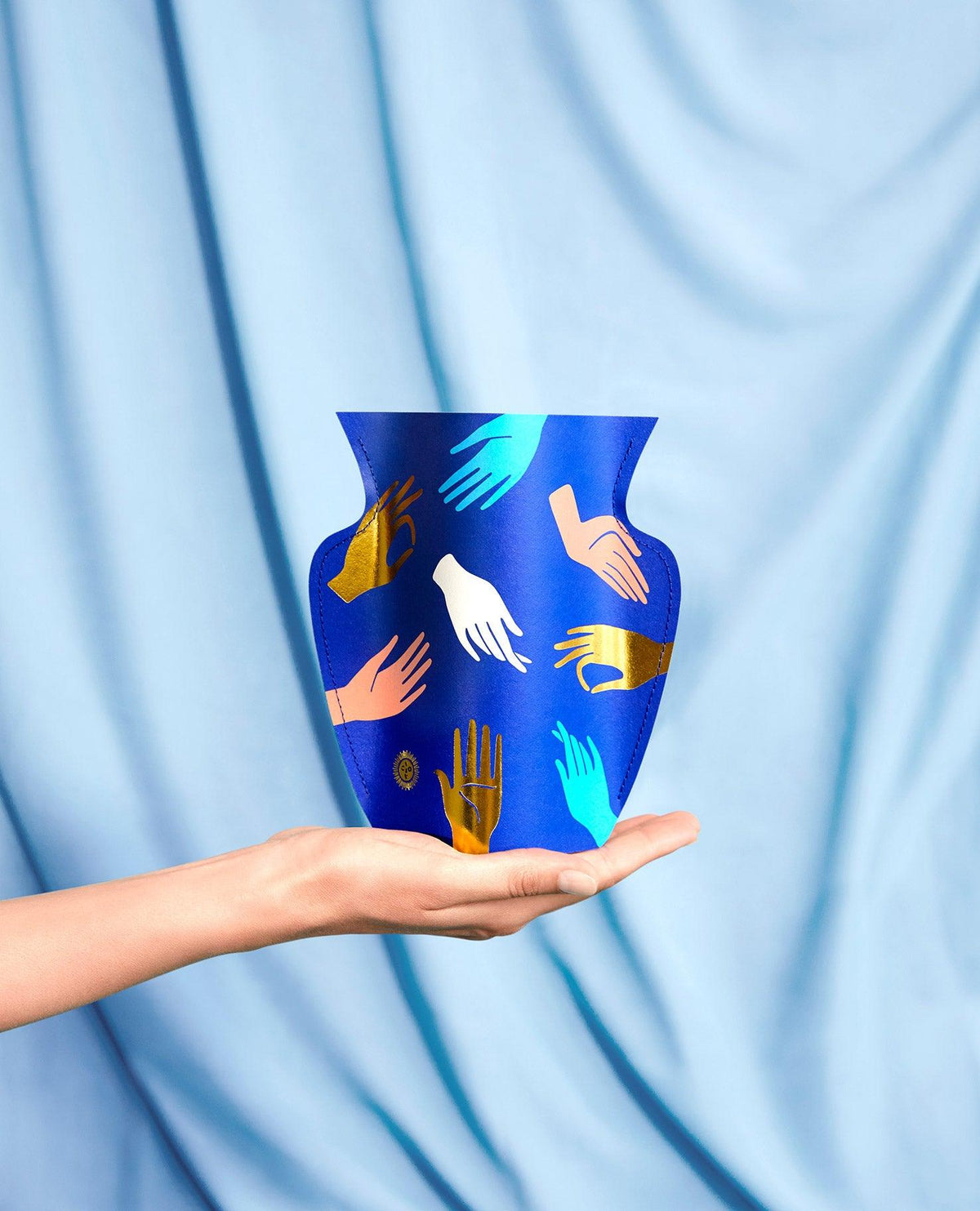 A hand holds the OCTAEVO Hamsa Paper Vase with colorful hand design, set against a light blue fabric backdrop.