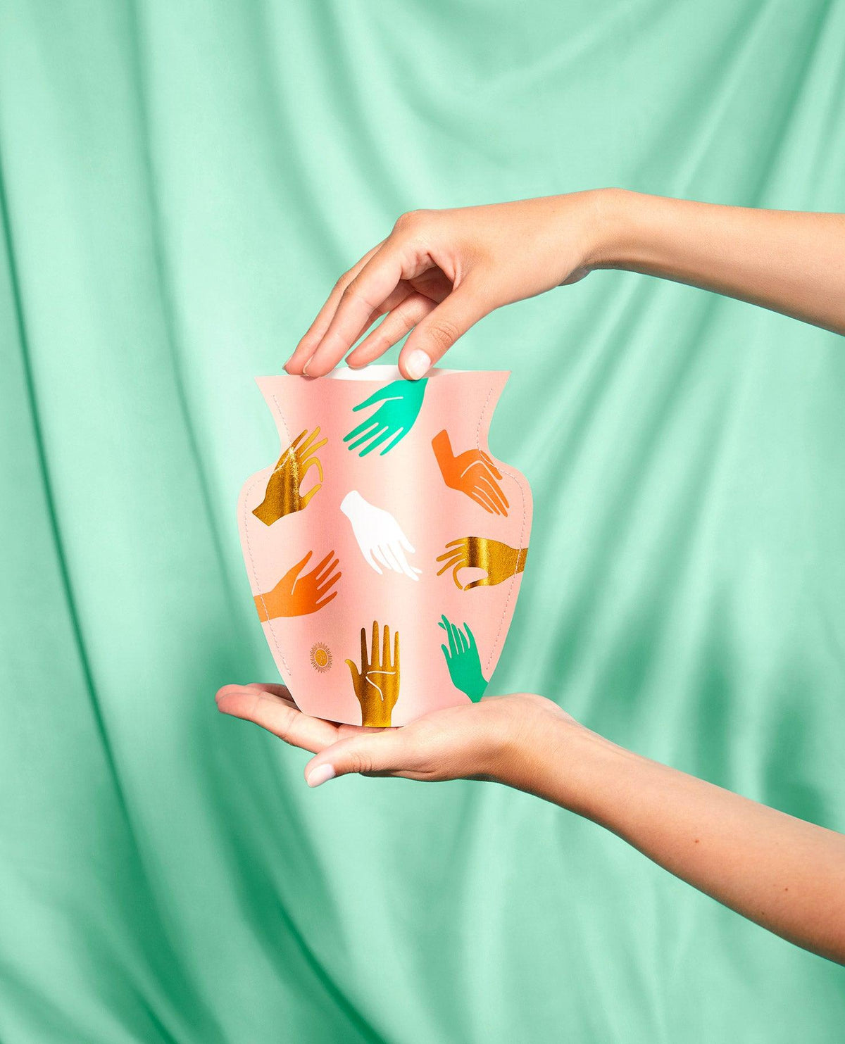 Two hands hold pink OCTAEVO Hamsa Paper Vase, featuring colorful gold hand motifs, against green draped fabric.