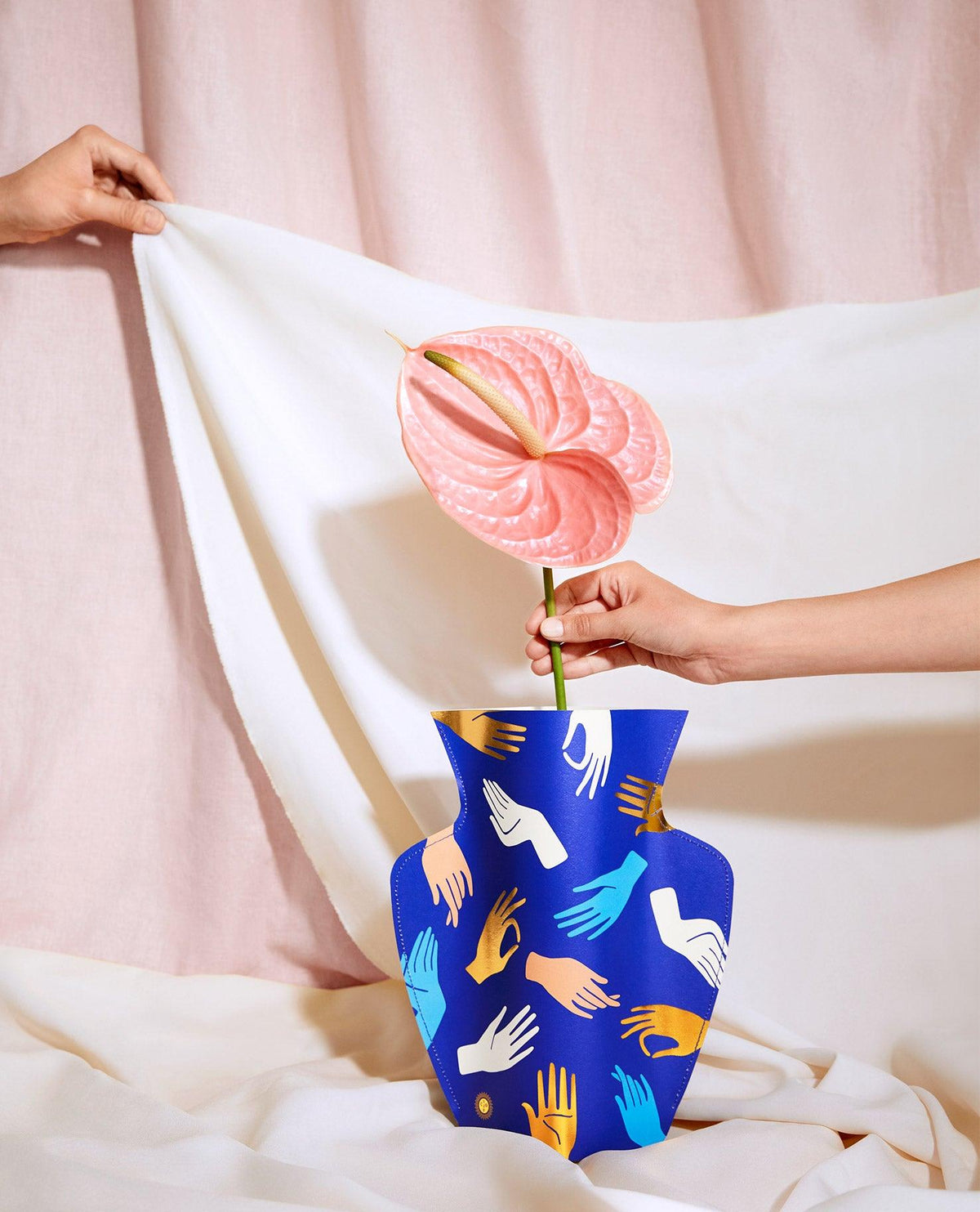 A hand holds a flower in the blue OCTAEVO Hamsa Paper Vase; another hand adjusts a white cloth backdrop.