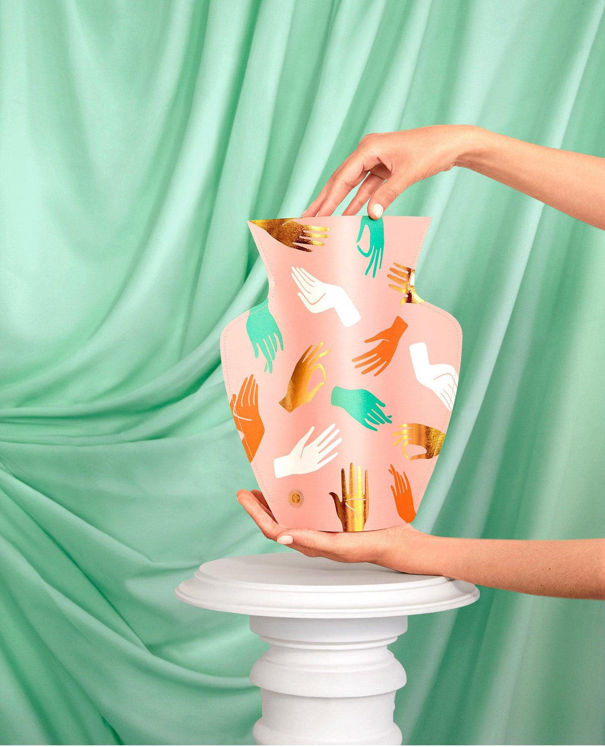 A hand holds the pink OCTAEVO Hamsa Paper Vase with colorful prints, set against a mint green draped fabric. 
