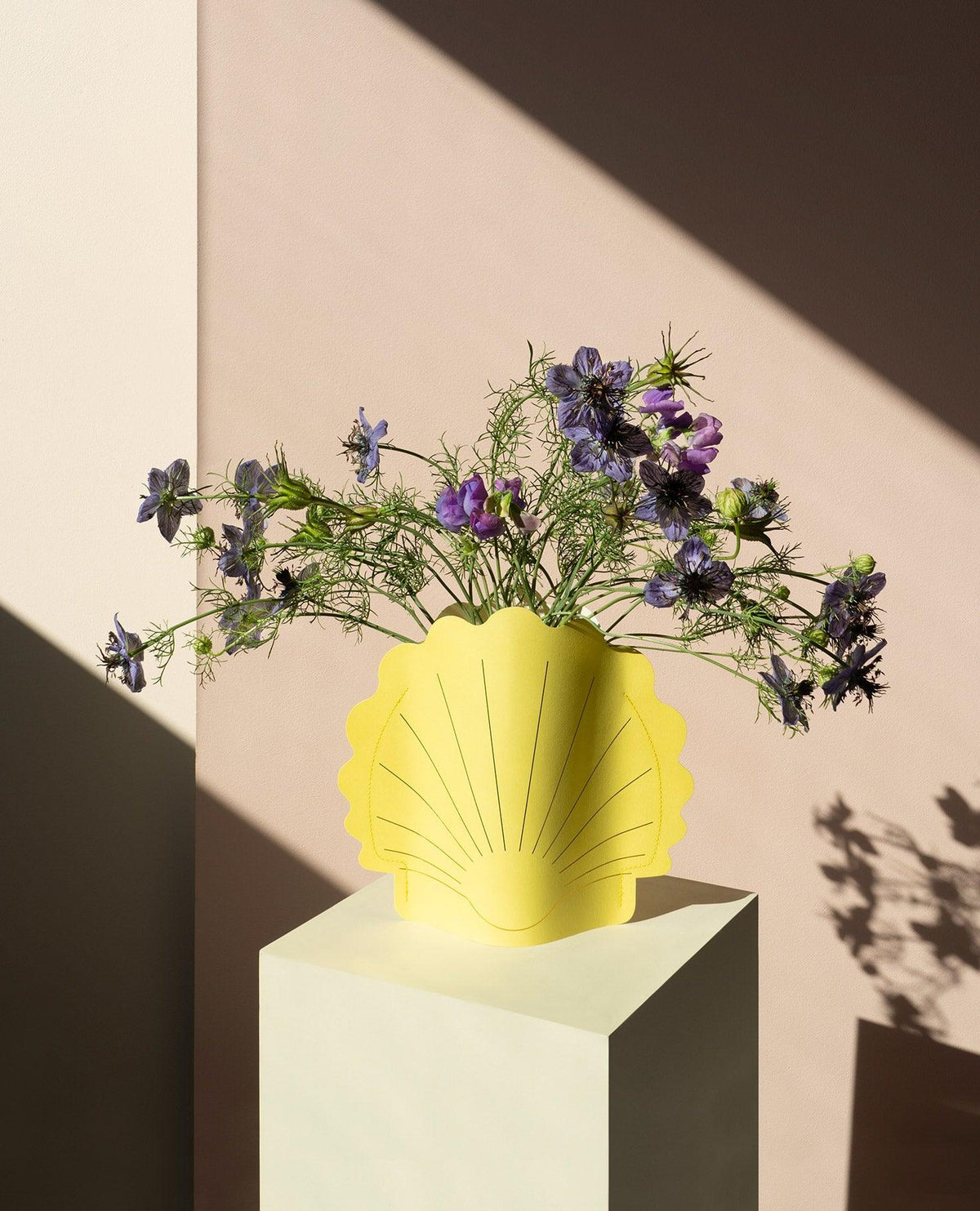 The Hera Paper Vase by OCTAEVO, conch-shaped and yellow, holds purple flowers on a sunlit white pedestal.