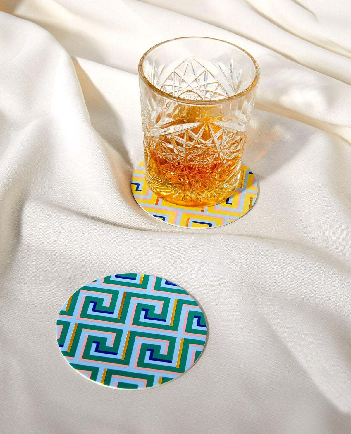 A glass of amber liquid rests on an OCTAEVO Icarus Cocktail Coaster, gold foil accents nearby on white fabric.