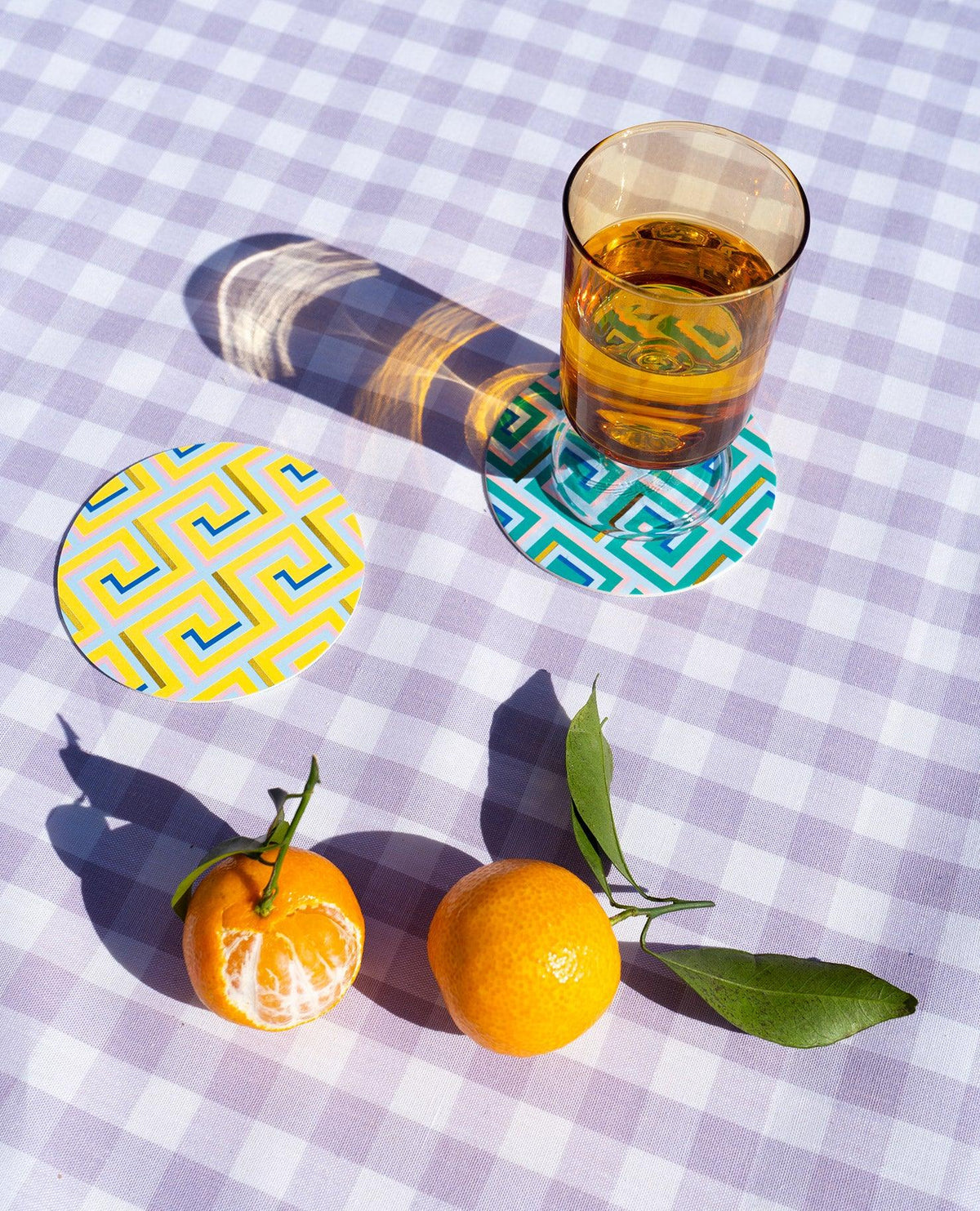 An amber glass, tangerines, and OCTAEVO Icarus Cocktail Coasters on a purple checked tablecloth.