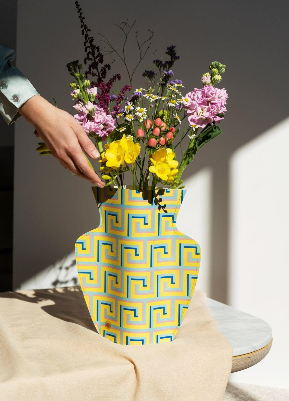 A hand arranges wildflowers in the yellow OCTAEVO Icarus Paper Vase with geometric patterns on a sunlit table.