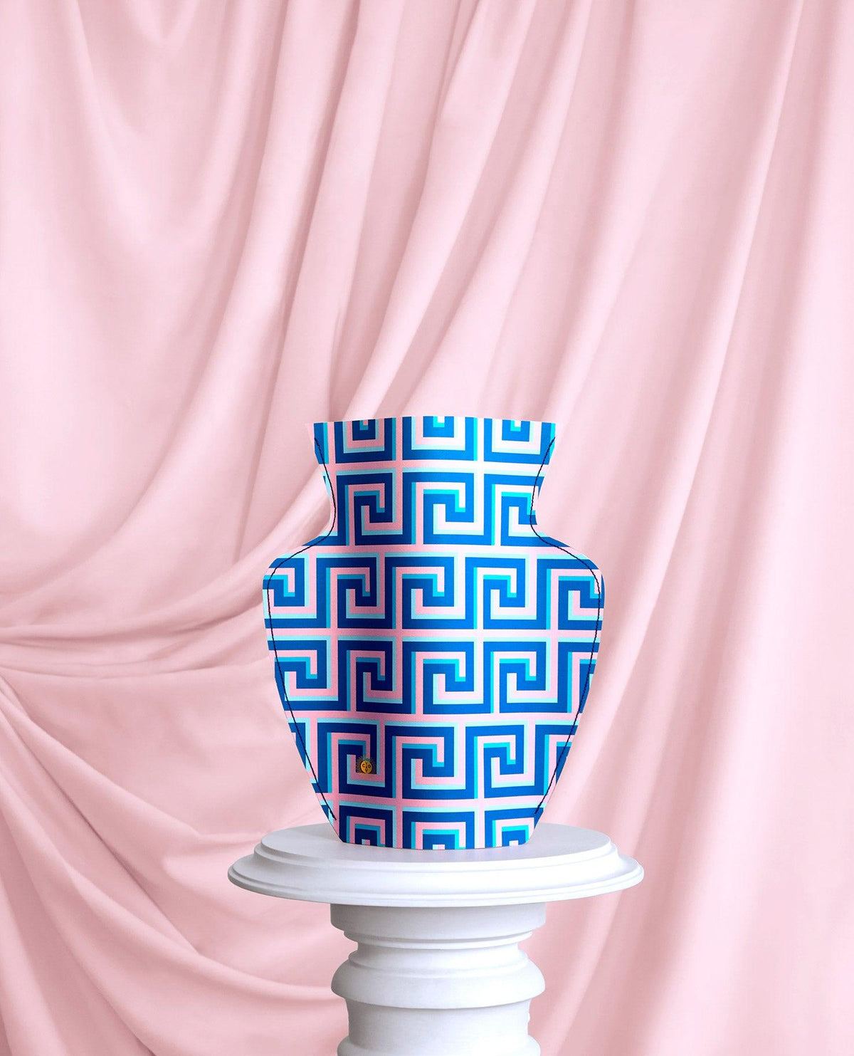 The OCTAEVO Icarus Paper Vase in blue and pink is displayed on a pedestal with a pink draped fabric backdrop.