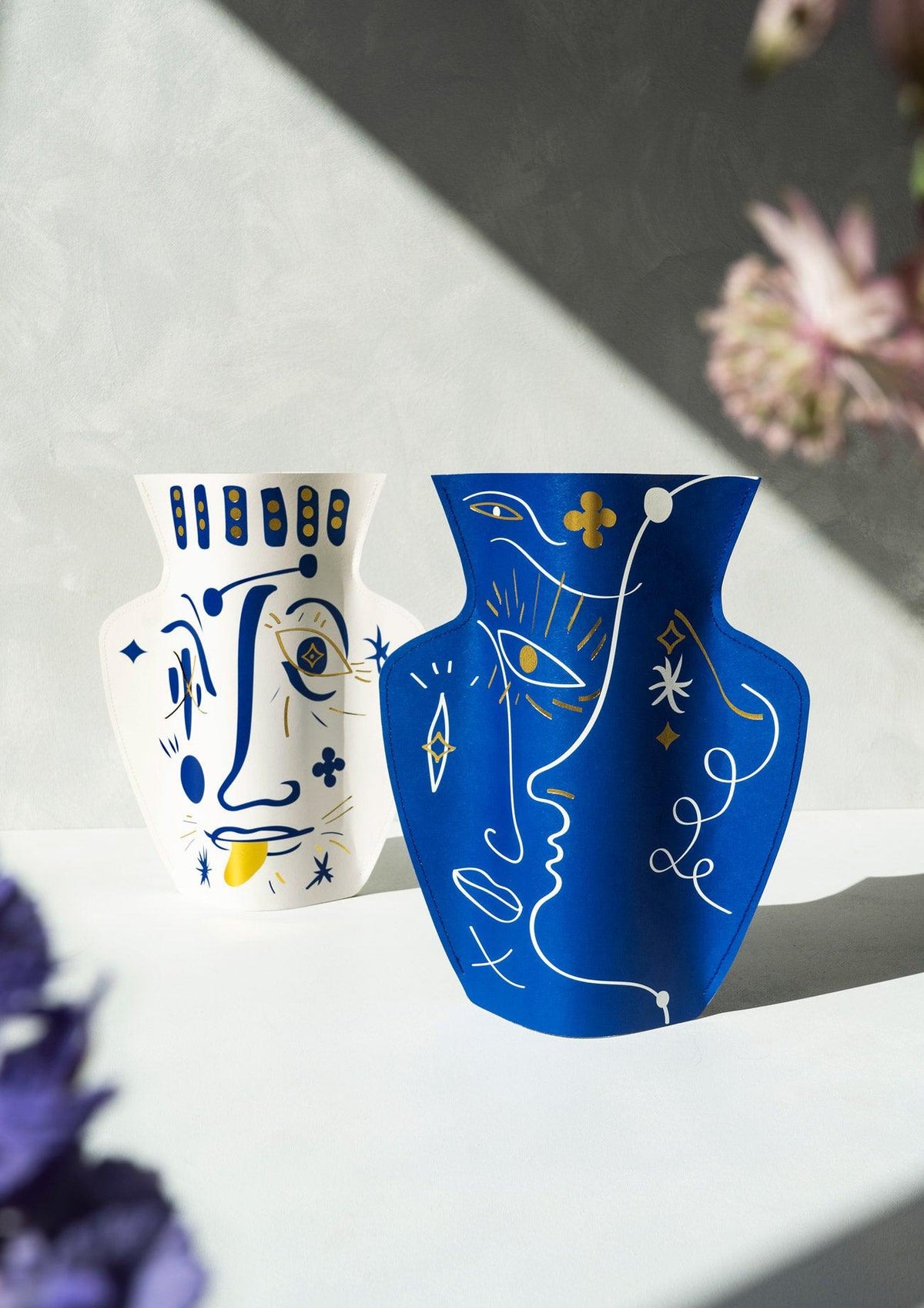 Two OCTAEVO Jaime Hayon Paper Vases in Vasage White sit sunlit with flowers nearby.