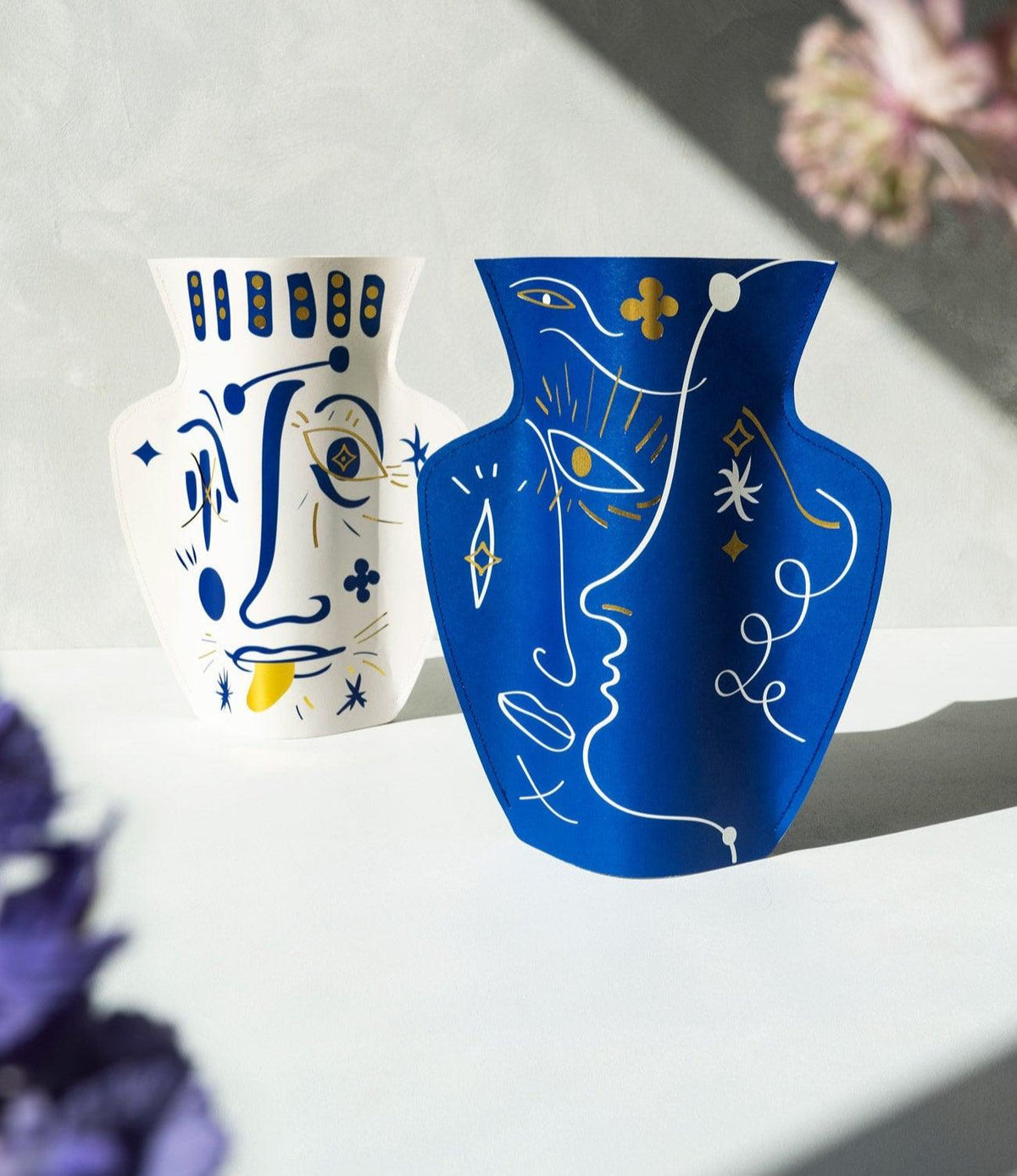 Two OCTAEVO Jaime Hayon Paper Vases in Vasage Blue with abstract faces are displayed on a sunlit surface.