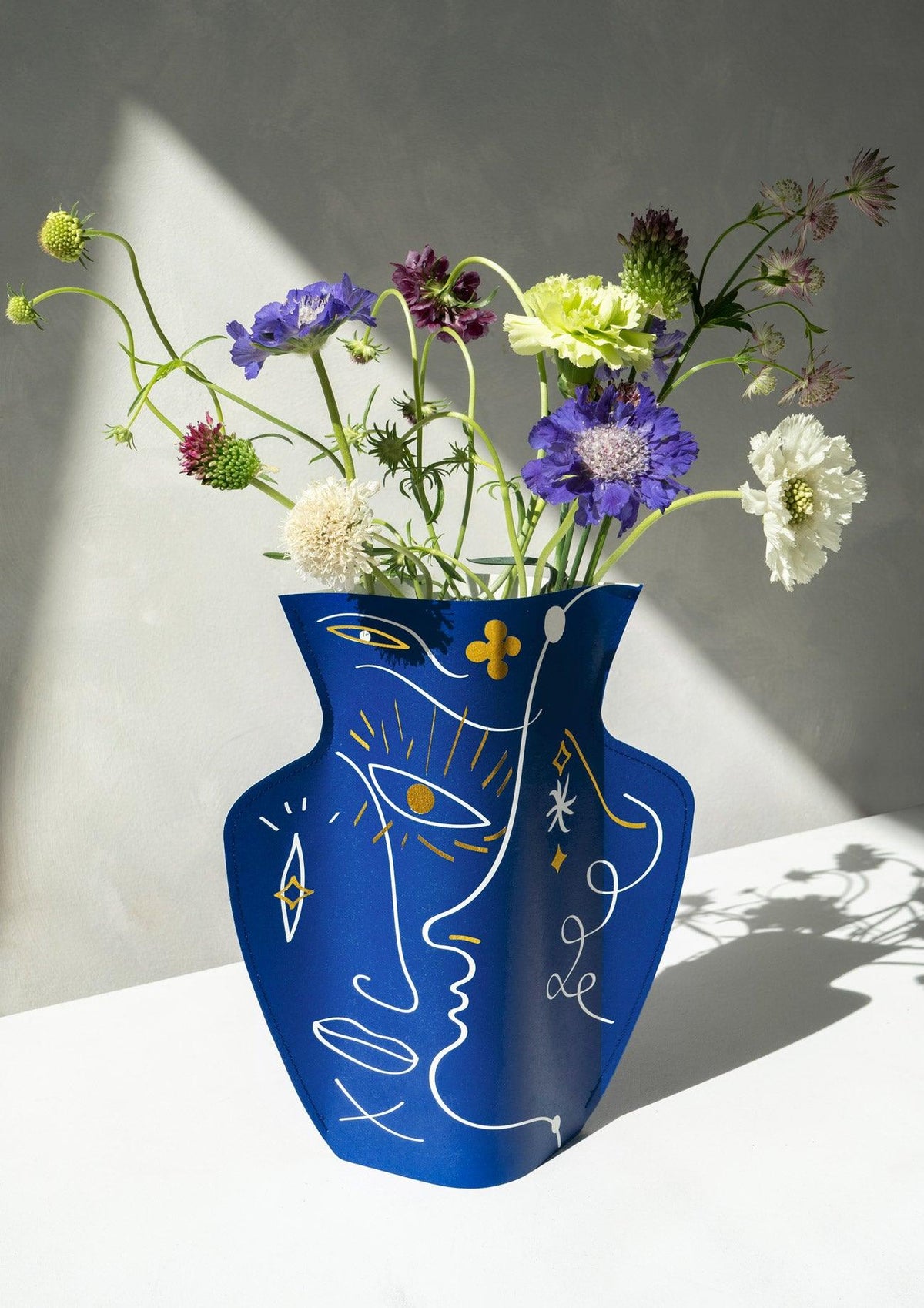 The OCTAEVO Jaime Hayon Paper Vase with abstract faces holds purple, white, and green flowers in sunlight.