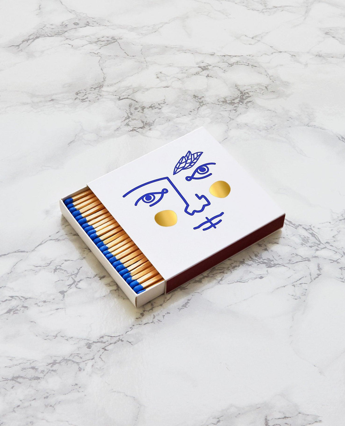 A box of Janus Matches by OCTAEVO features blue tips, a minimalist face design, and gold foil accents on marble.