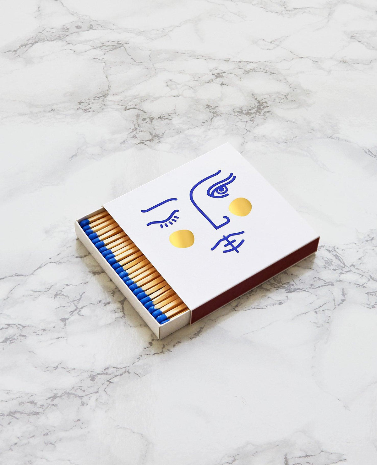 Allumettes Janus par OCTAEVO ornées d'un visage minimaliste en bleu et dorure à chaud, sur du marbre.