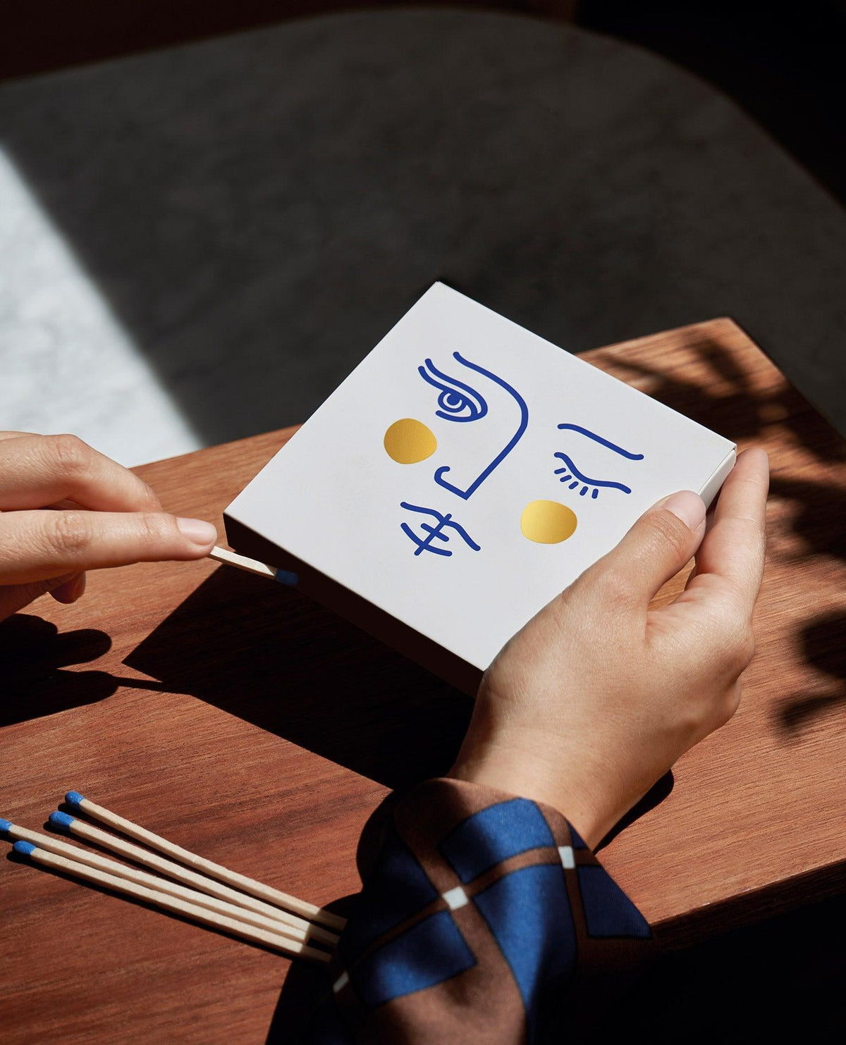 Hands holding a Janus Matches box by OCTAEVO with a minimalist face design and gold foil, beside matches on wood.