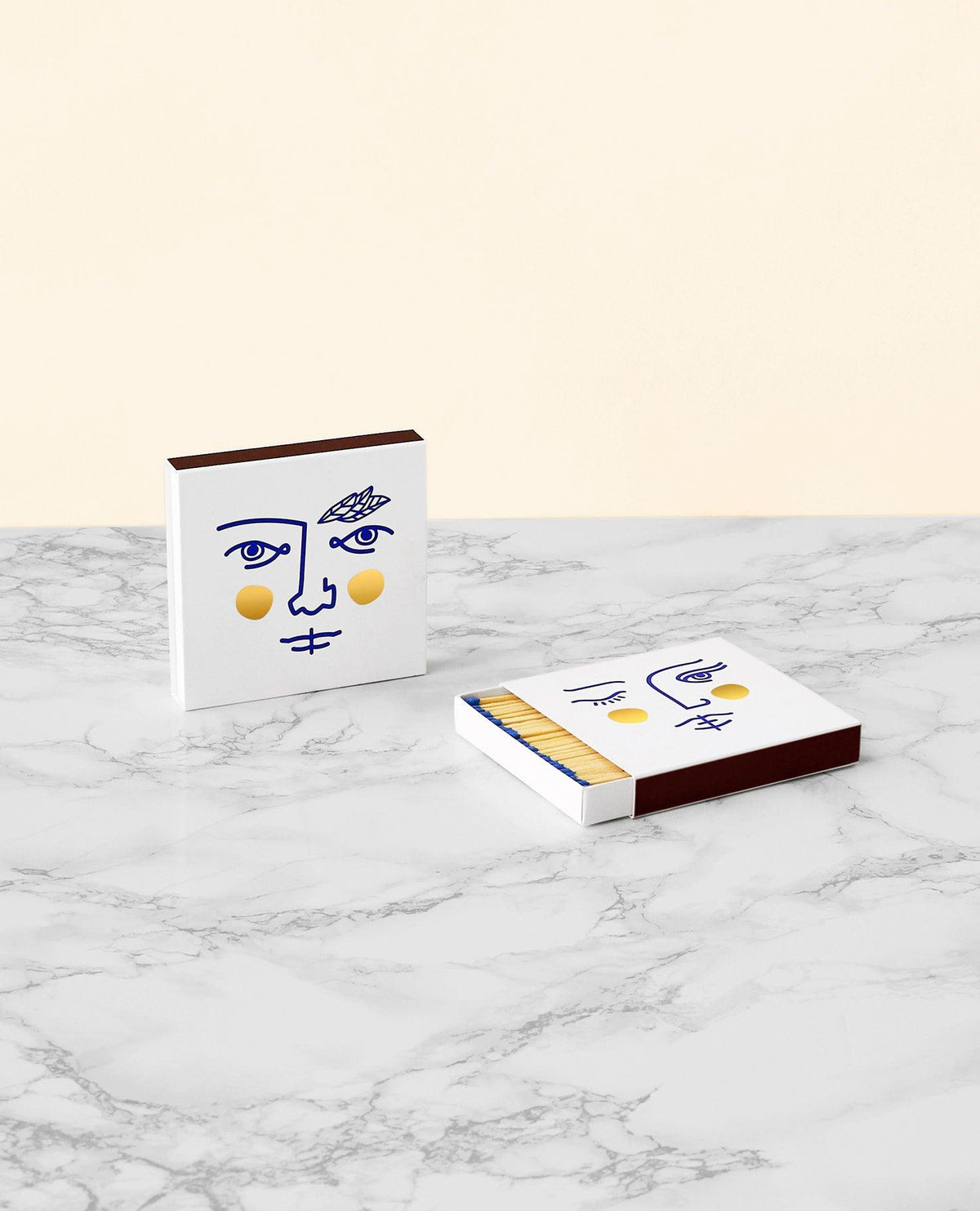 Deux boîtes d'allumettes Janus par OCTAEVO aux visages minimalistes et à la dorure à chaud, sur du marbre. Une boîte est ouverte et laisse voir les allumettes.
