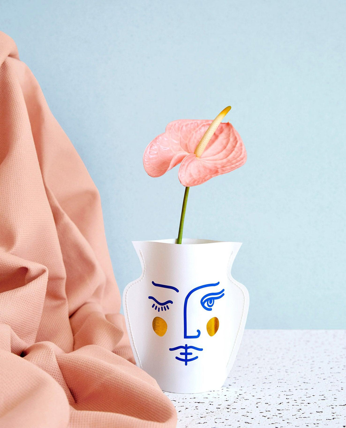 A pink anthurium in OCTAEVOs Janus Paper Vase with a blue face design, set on a pastel background.