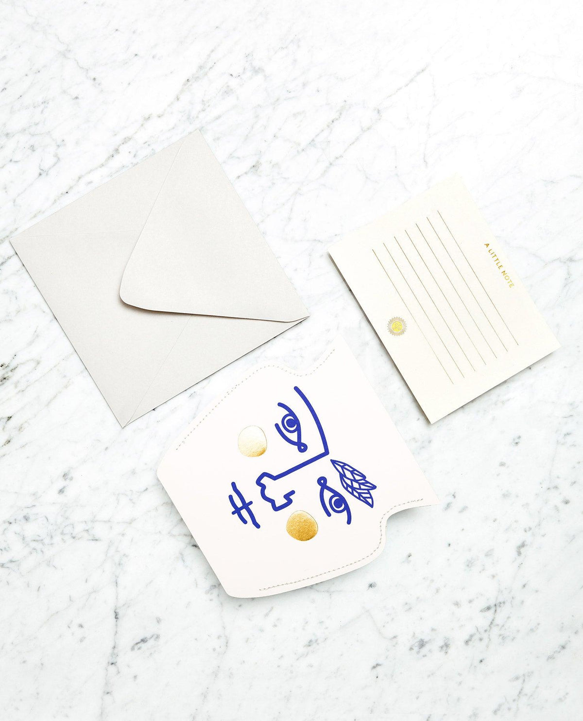 A blue and gold abstract face paper vase, envelope, and OCTAEVO Janus Mini Paper Vase are displayed on a marble surface.