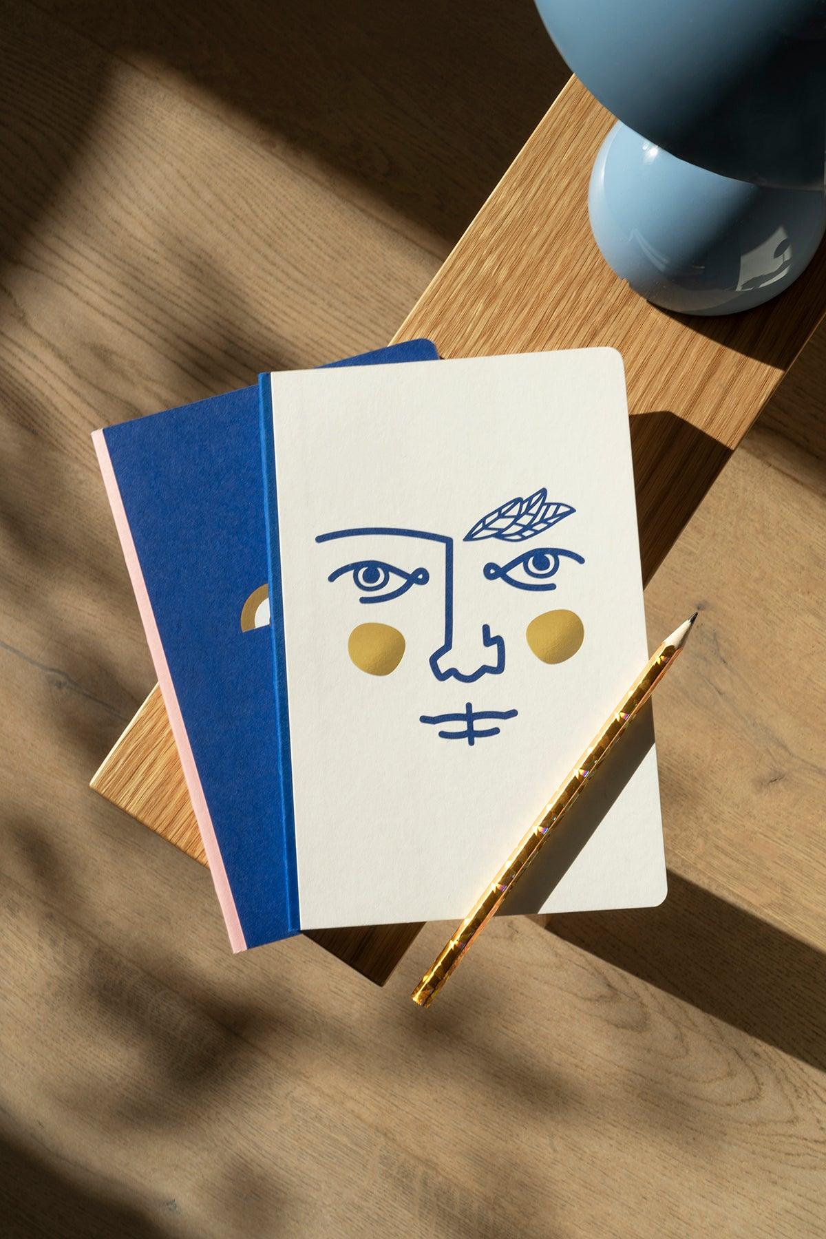 Two Octaevo Janus Notes A5 luxury notebooks and a gold pen with lay-flat binding on a sunlit wooden surface.