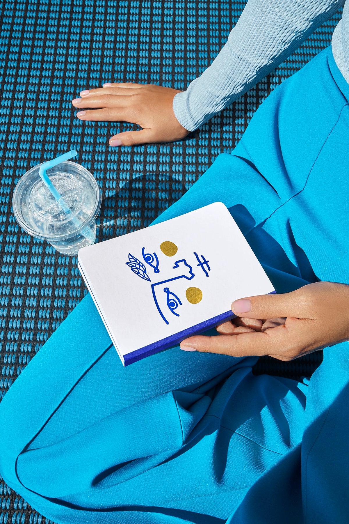Person in blue pants holds an Octaevo Janus Notes A6 notebook with abstract face, beside a clear drink on blue woven surface.