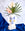 The OCTAEVO Janus Paper Vase with a blue abstract face holds palm leaves and anthuriums on blue fabric.