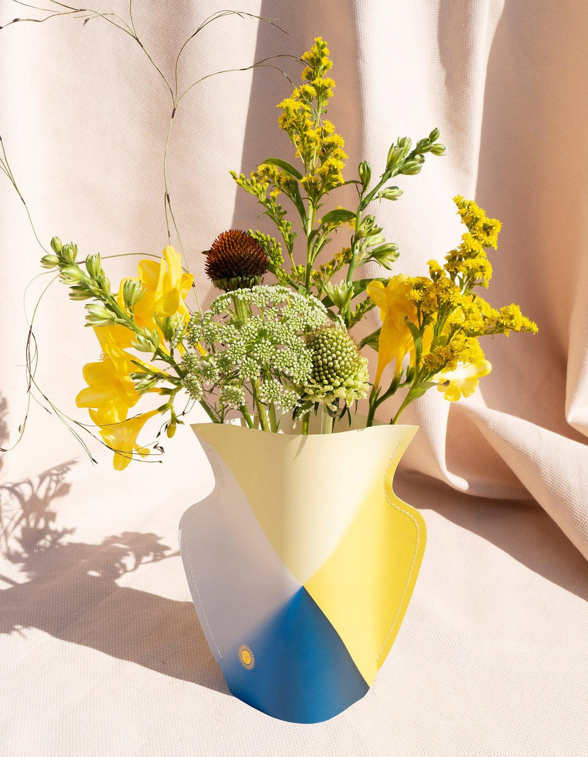 The OCTAEVO Laguna Paper Vase, in yellow and white, adds Mediterranean flair to blooms against a beige fabric backdrop.