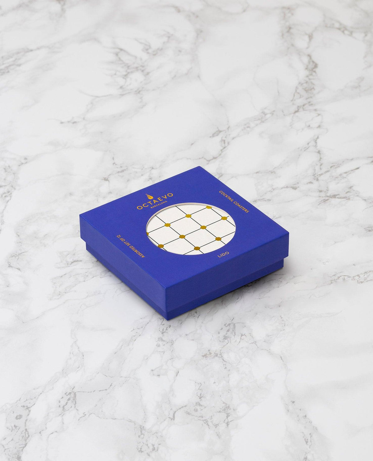 The Lido Cocktail Coasters by OCTAEVO, in a blue square box with gold foil accents, on white marble.