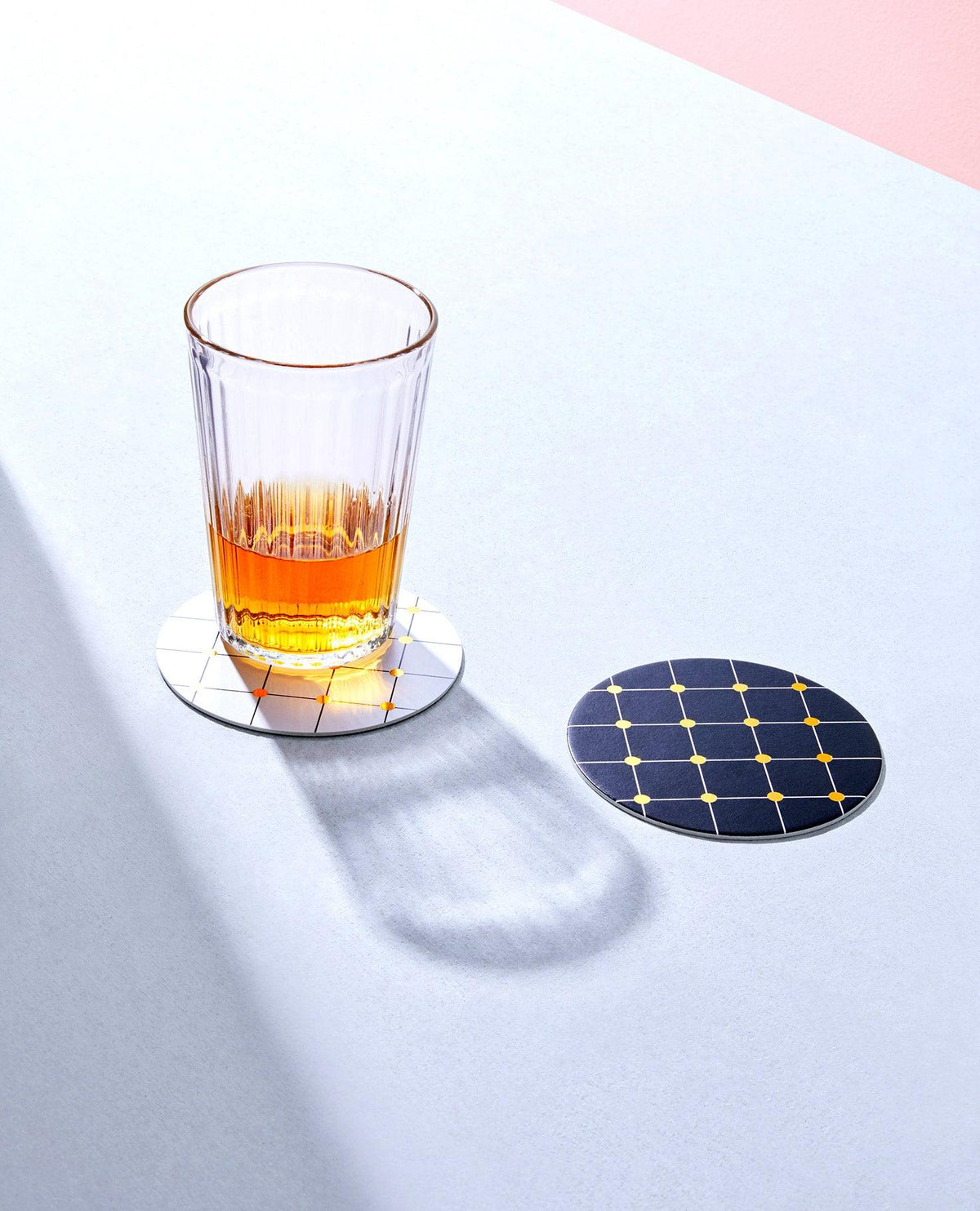 A glass of amber liquid rests on OCTAEVOs gold-foiled Lido Cocktail Coasters, on a light surface.