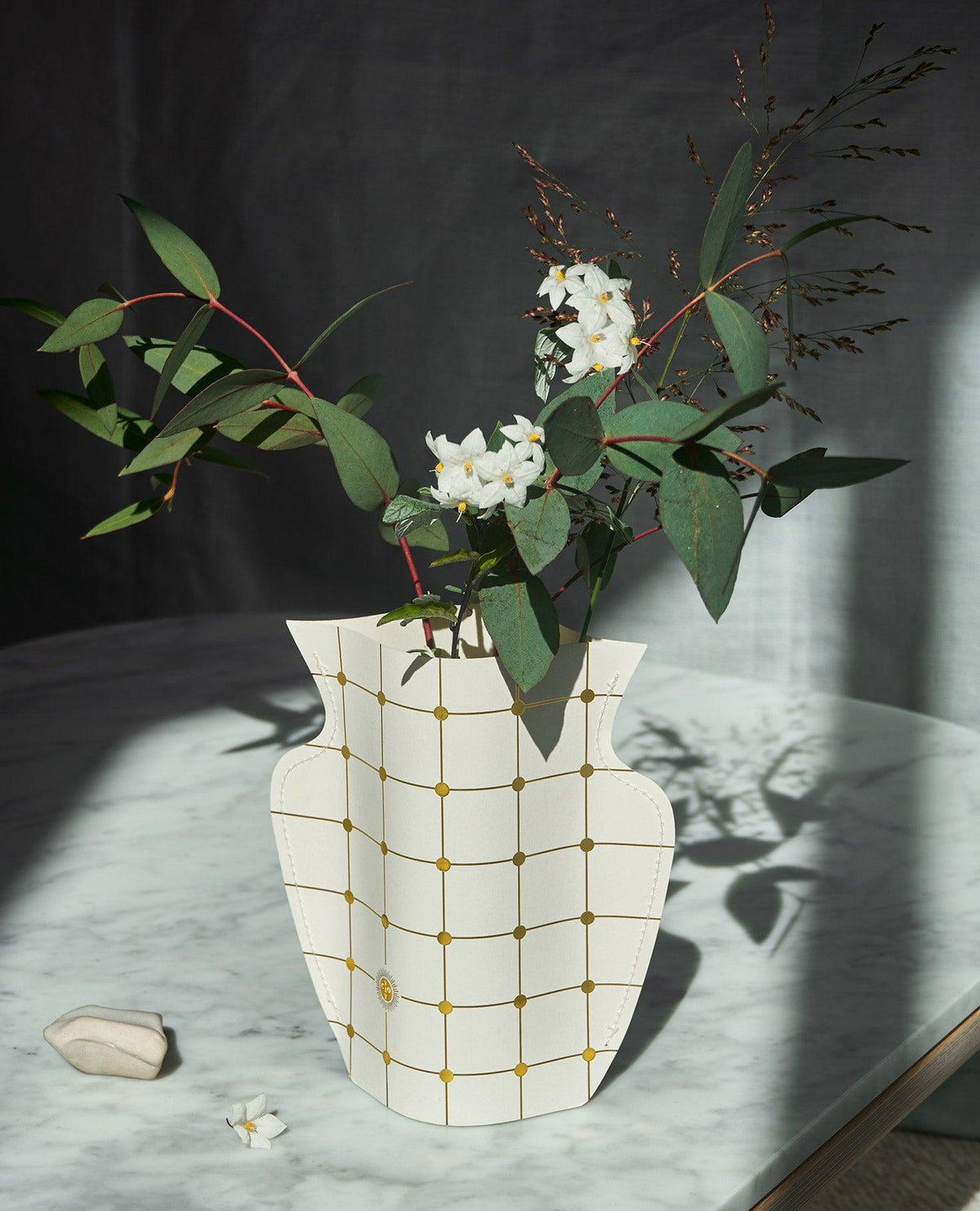 The OCTAEVO Lido Mini Paper Vase with grid pattern holding greenery and white flowers on a sunlit marble table.