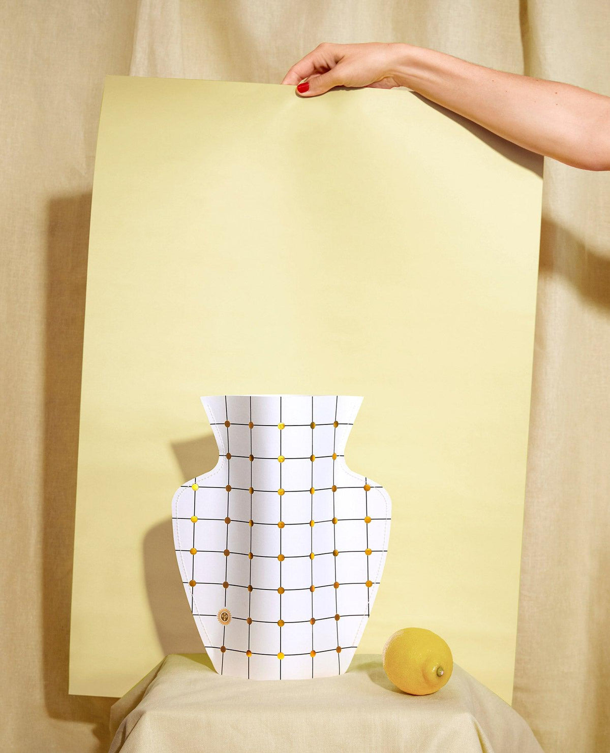 A hand holds a large sheet behind a white OCTAEVO Lido Paper Vase and a lemon. 