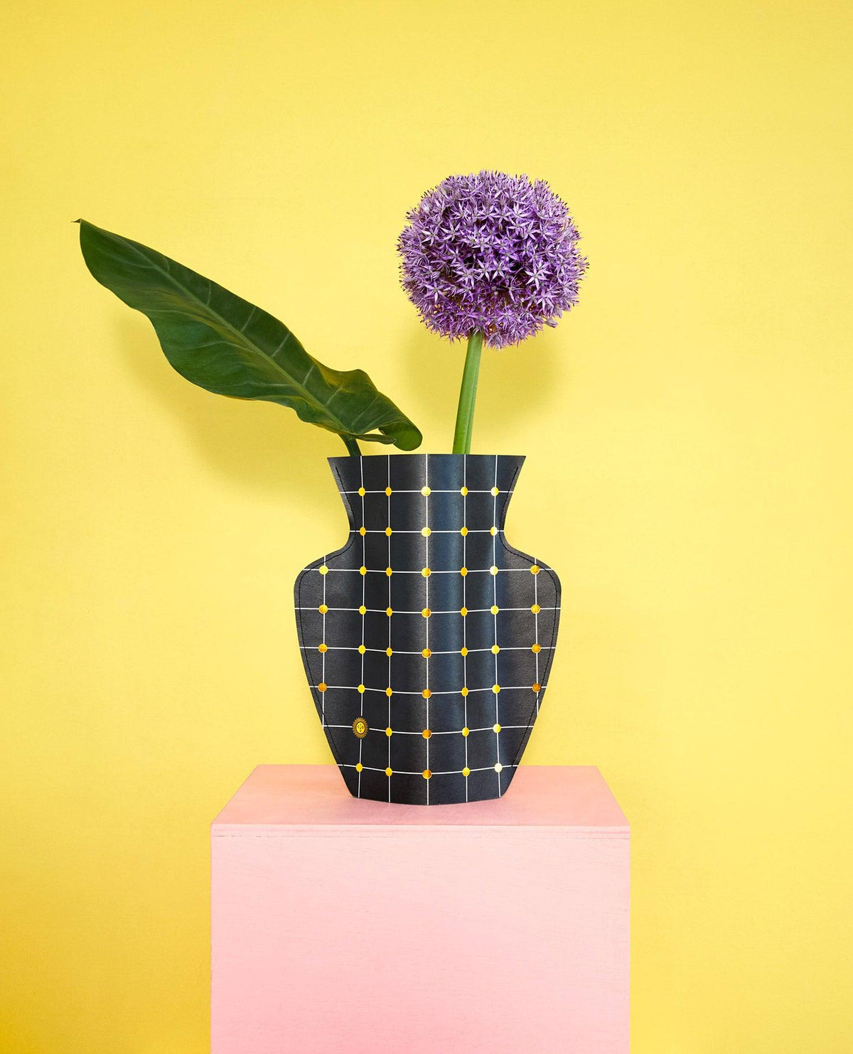 The black Lido Paper Vase by OCTAEVO features a gold grid, displaying a green leaf and purple flower against a yellow wall.