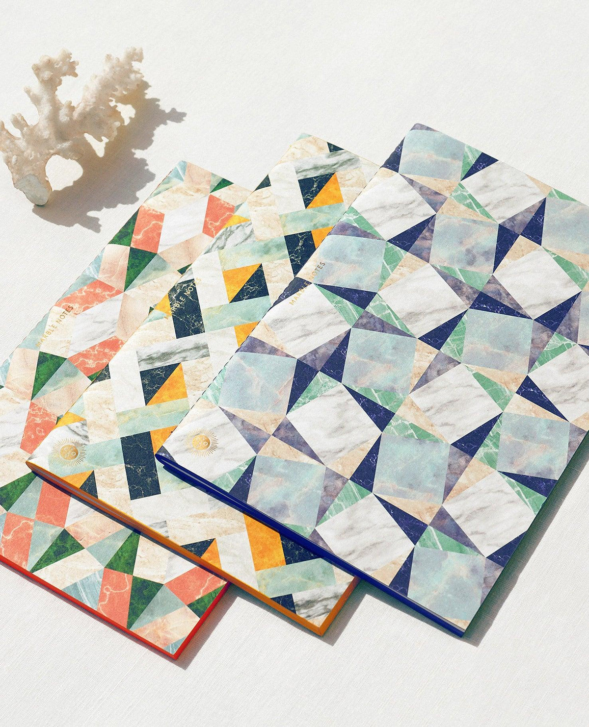 Three OCTAEVO Marble Notes notebooks with colorful geometric covers rest next to white coral on a bright surface.