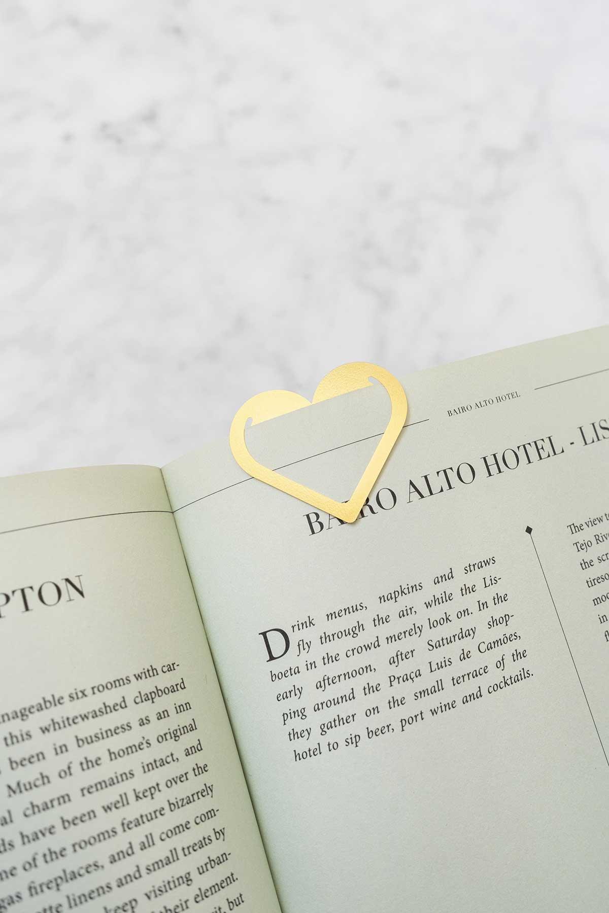 A heart-shaped clip in brass gold by OCTAEVO attached to a mint book page on a marble surface.